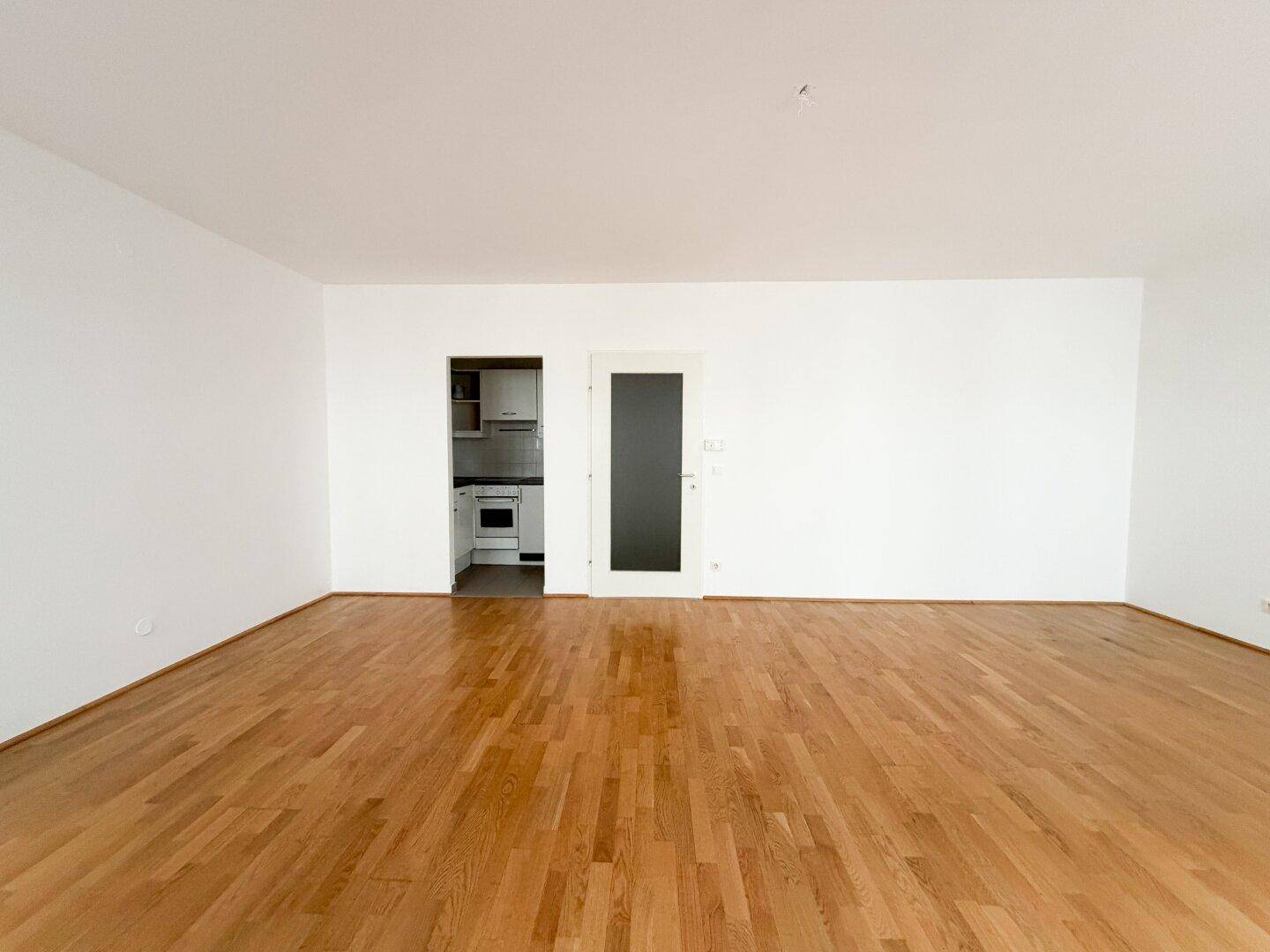 Geräumiges Zimmer mit Parkettboden und Zugang zu einer kleinen Küche, ideal für offenes Wohnen.