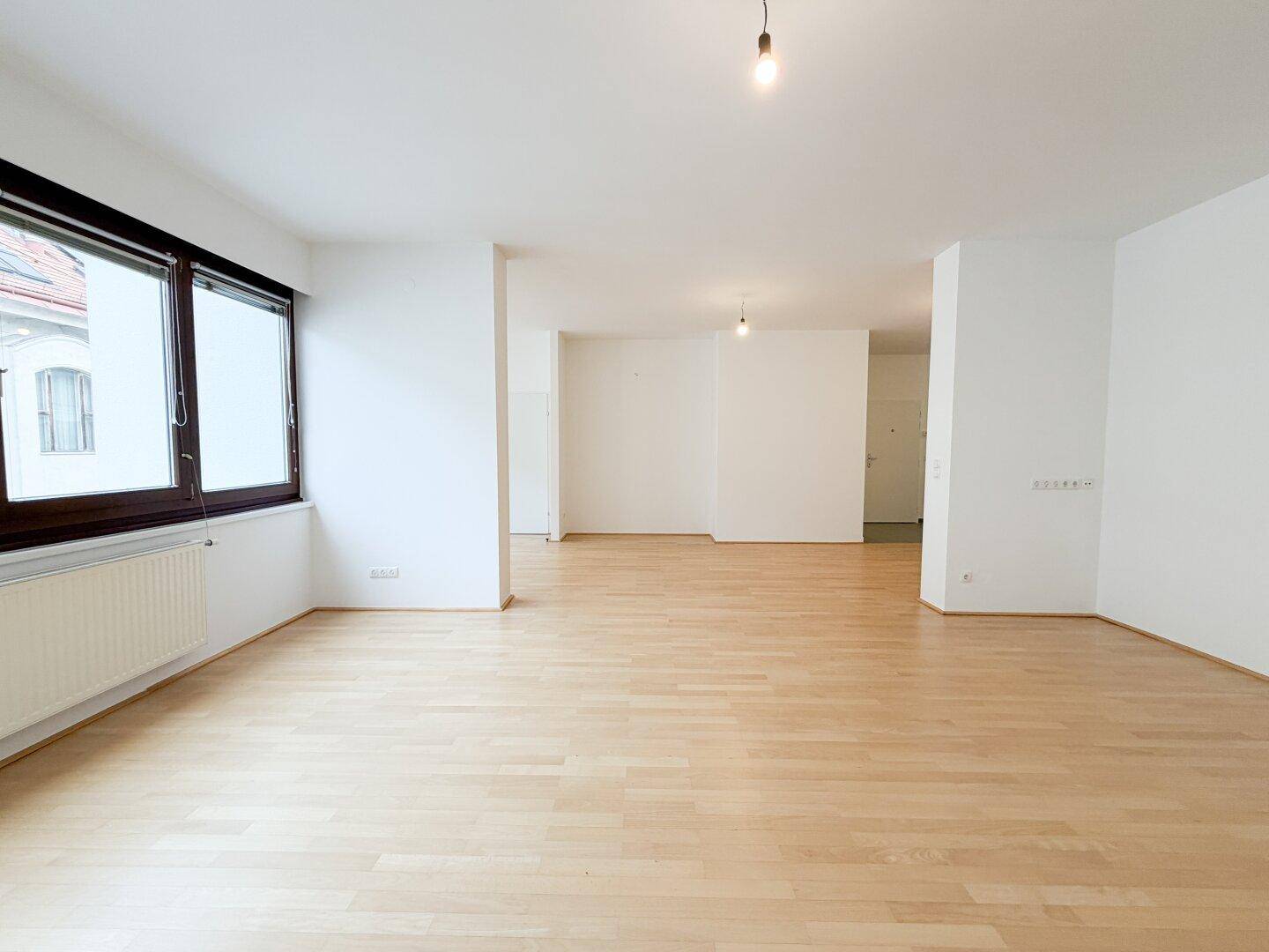 Heller, leerer Wohnbereich mit Parkettboden und Blick auf die umliegenden Gebäude.