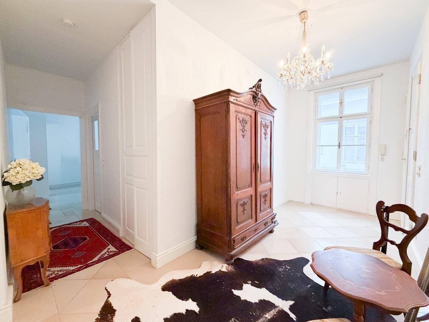 Heller Flur mit antikem Holzschrank, kleinem Tisch und Blick in einen weiteren Raum.