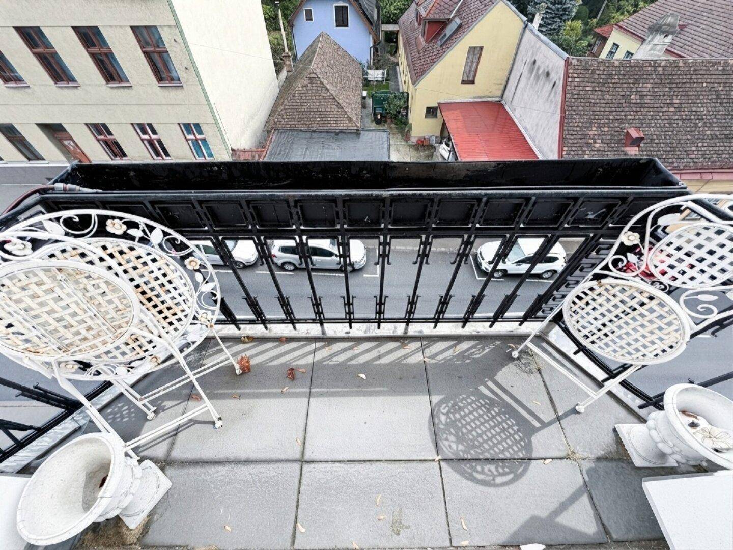 Balkon mit schmiedeeisernen Stühlen und Blick auf die darunterliegende Straße mit parkenden Autos.