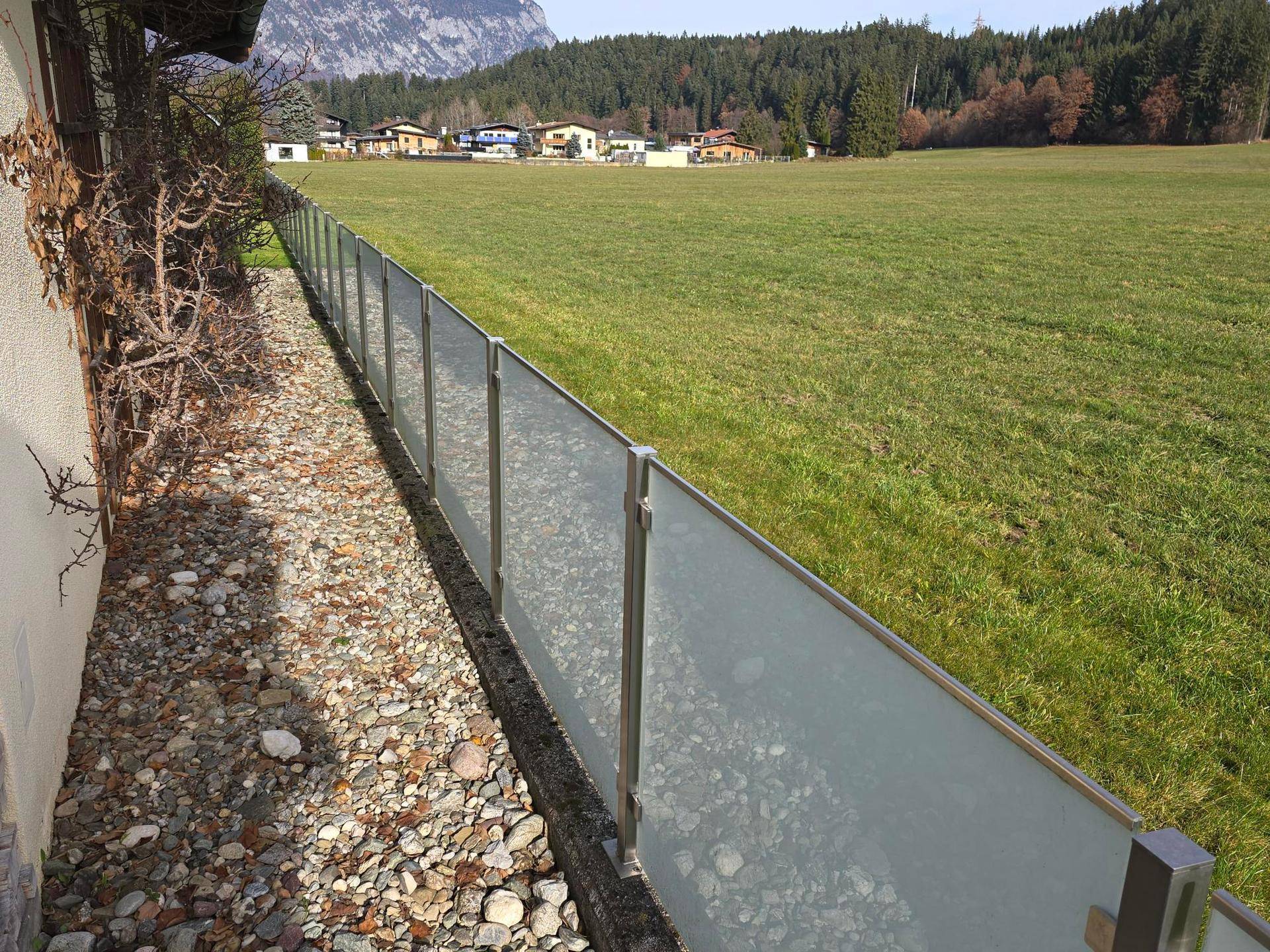 Gartenrand mit moderner Glasabtrennung und weitem Blick über grüne Felder und Berge.