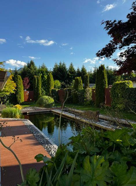 Detailansicht der Terrasse mit Teich, üppiger Bepflanzung und hohen Bäumen im Hintergrund.