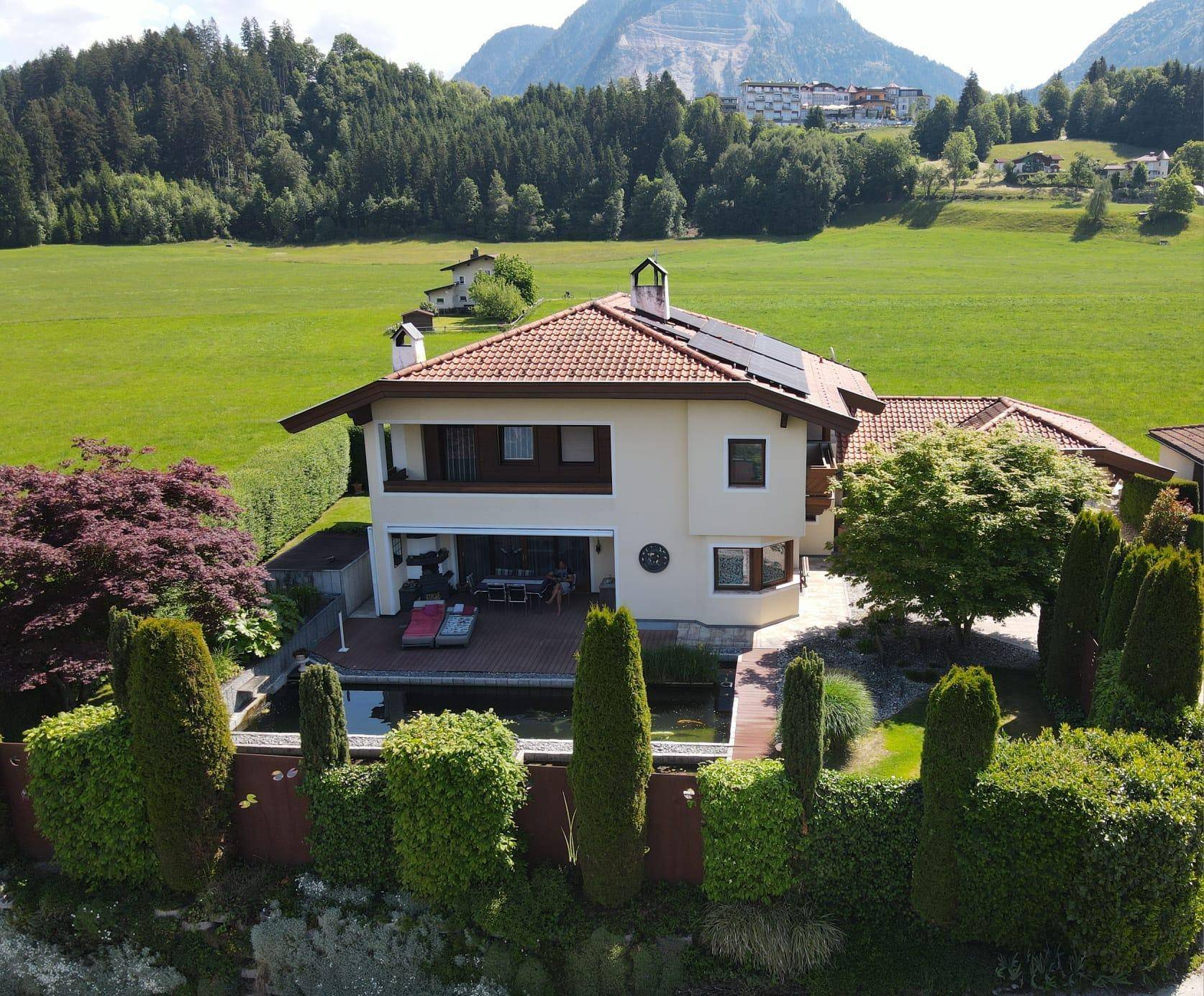 Luftaufnahme eines Hauses mit gepflegtem Garten, Teich, Terrasse und Bergblick in grüner Umgebung.