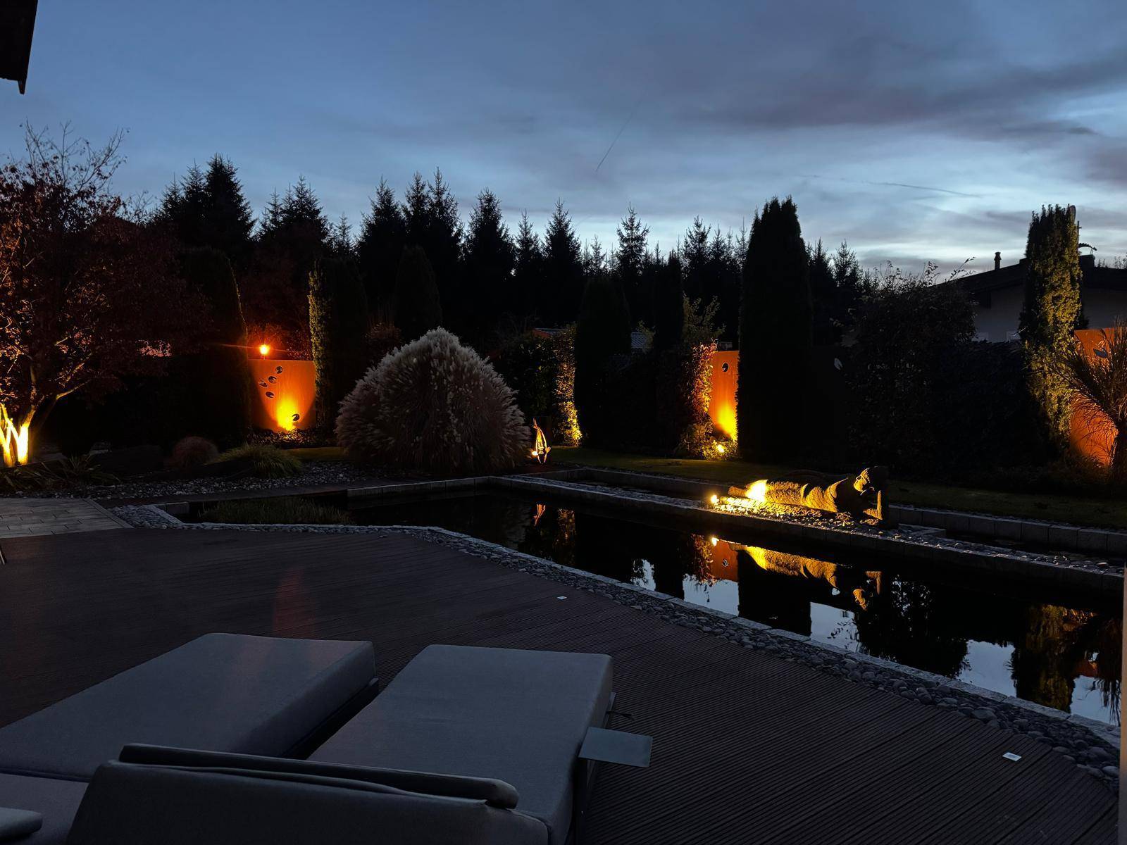 Abendliche Terrasse mit beleuchtetem Teich, Garten und bequemen Liegestühlen für entspannte Stunden.