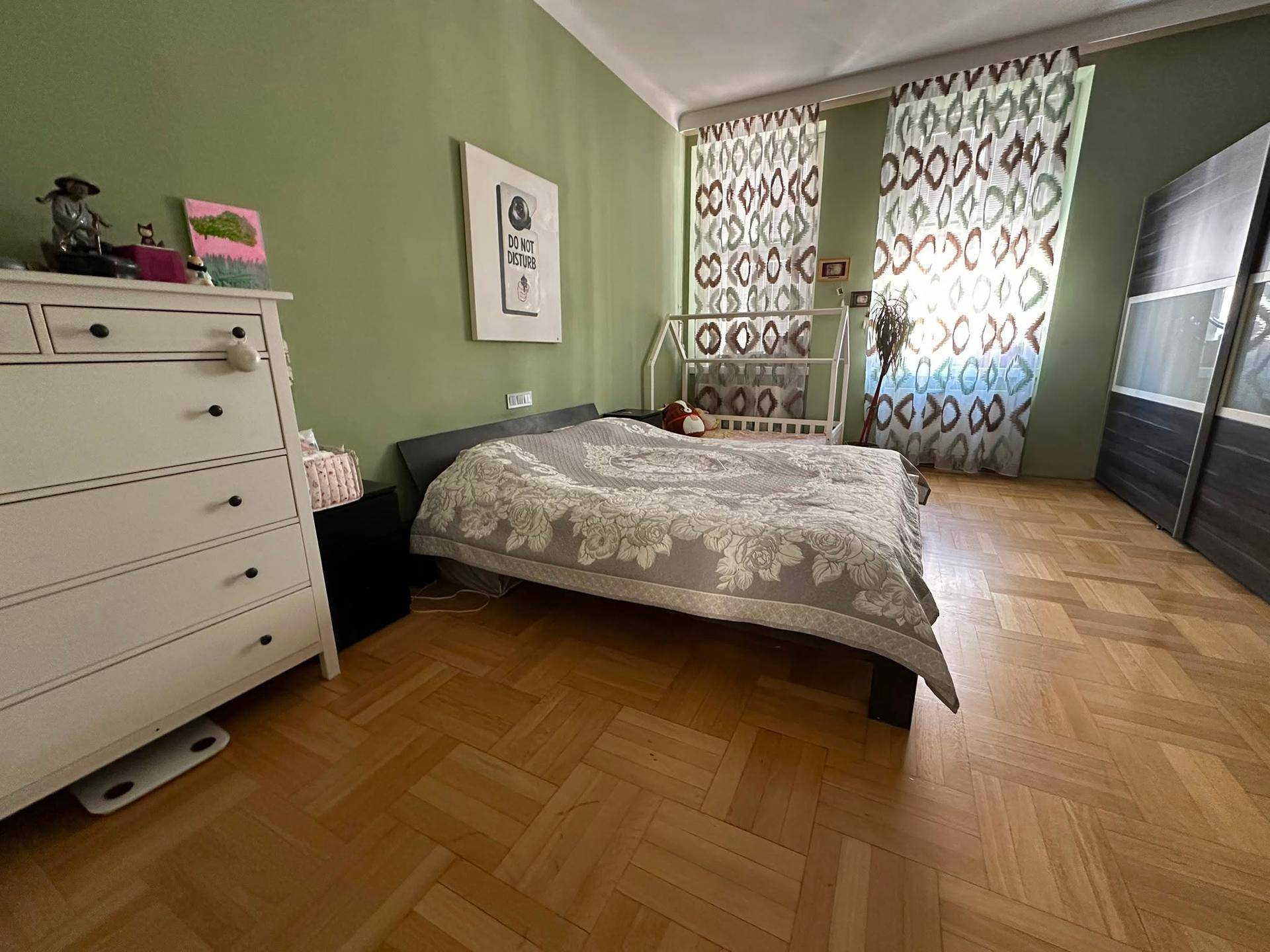 Helles Schlafzimmer mit Kommode, Doppelbett und gemusterten Vorhängen.