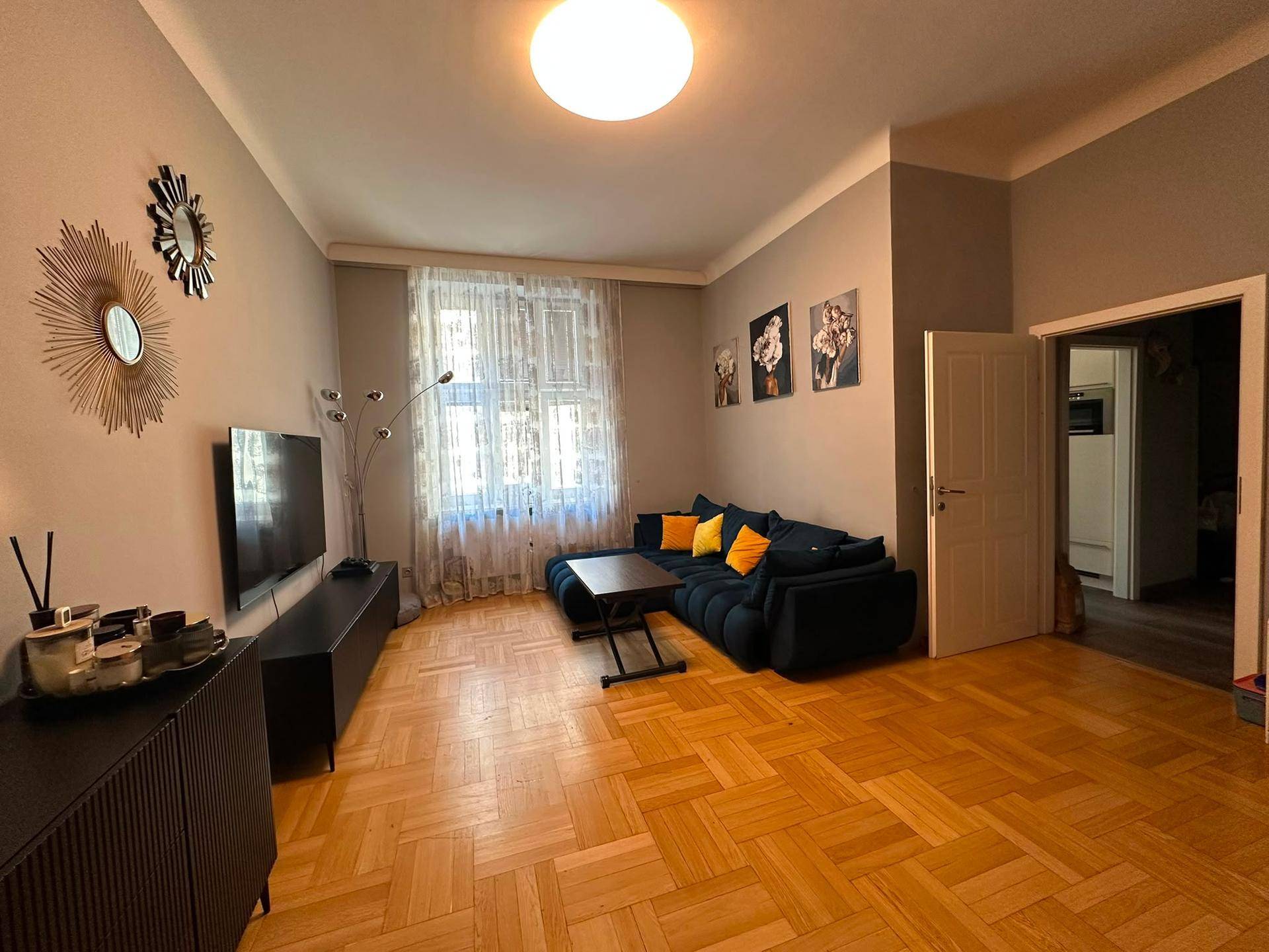 Geräumiges Wohnzimmer mit dunklem Sofa, Fernseher und hellem Fischgrätparkettboden.