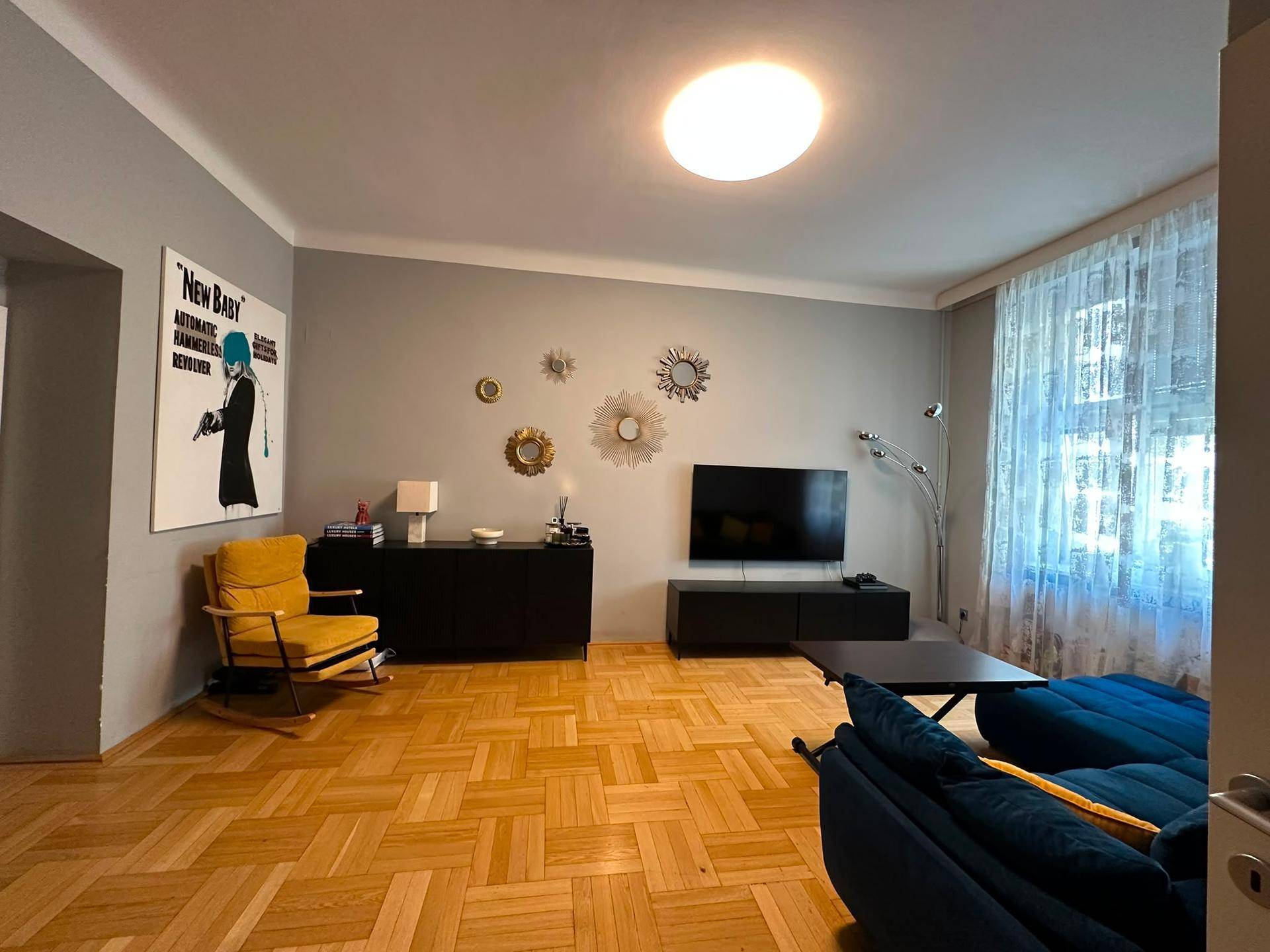 Geräumiges Wohnzimmer mit dunklem Sofa, Fernseher und hellem Fischgrätparkettboden.
