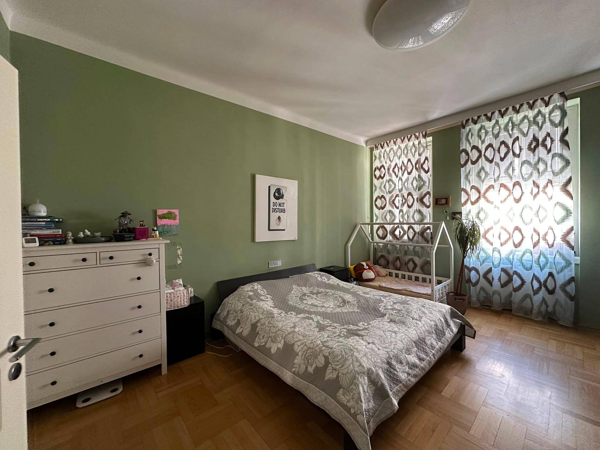 Helles Schlafzimmer mit Kommode, Doppelbett und gemusterten Vorhängen.