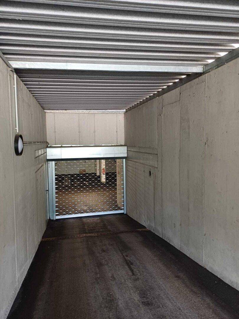 Innenansicht einer Tiefgarage mit Betonwänden und Rolltor am Ende des Ganges.