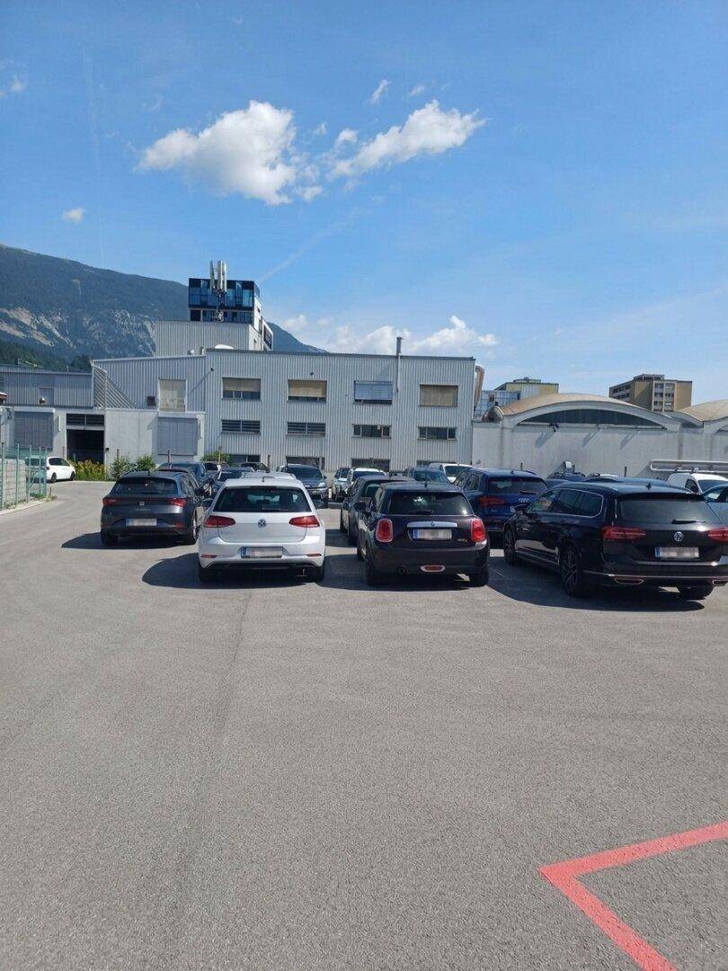 Großer Parkplatz mit zahlreichen Autos vor einem Industriegebäude und einer Berglandschaft unter blauem Himmel.