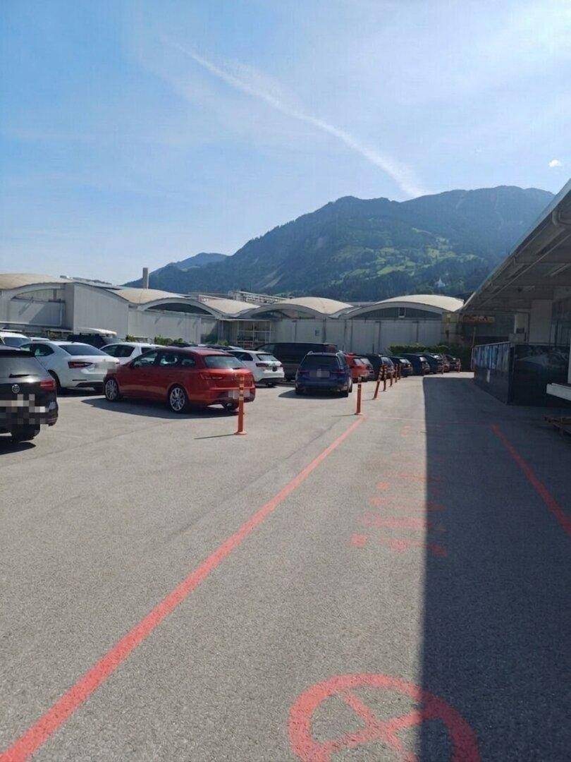 Ausgedehnter Parkplatz mit parkenden Fahrzeugen entlang einer Fahrspur, umgeben von Industriegebäuden und Bergen.
