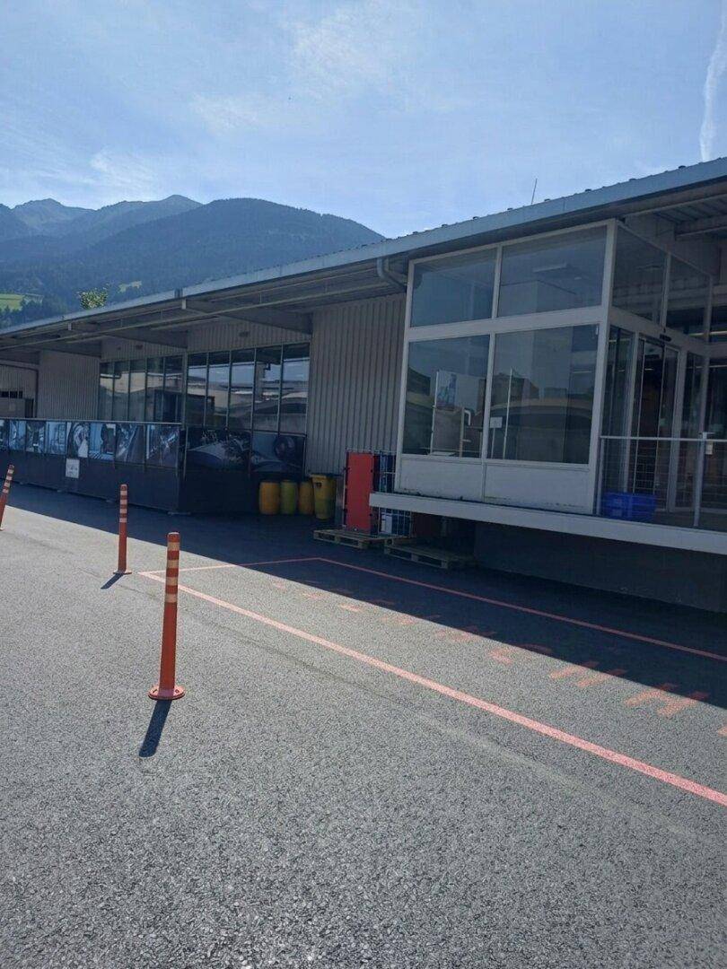 Ansicht der Gebäudefassade mit großflächigen Fenstern und einem Parkplatz im Vordergrund, umgeben von Bergen.