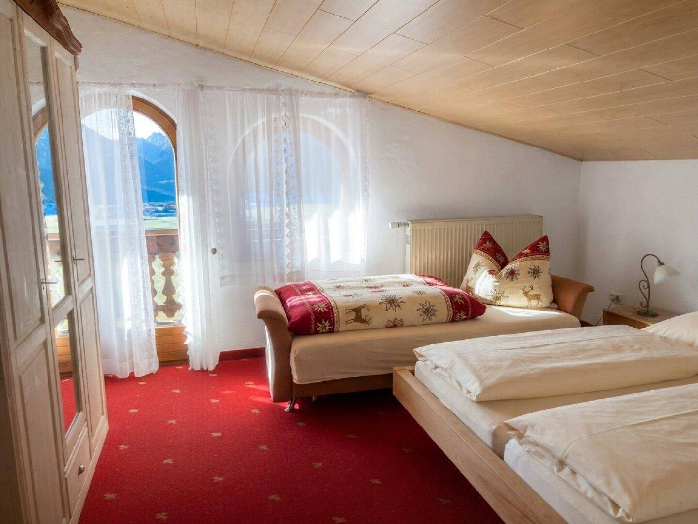 Helles Schlafzimmer im Dachgeschoss mit zwei Betten, rotem Teppich und Zugang zum Balkon.