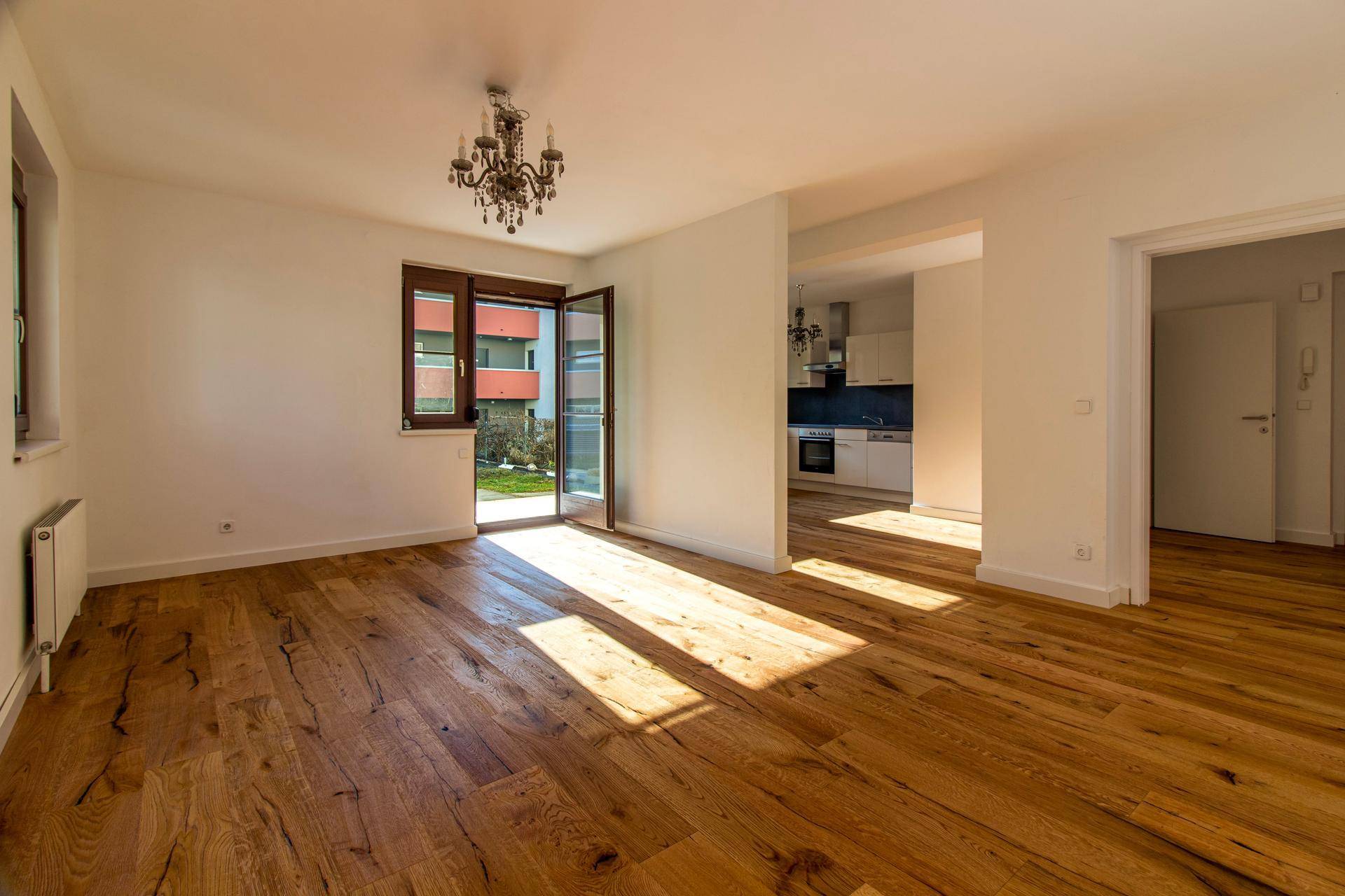 Heller Wohnraum mit Holzboden, Kronleuchter und offenem Übergang zur angrenzenden Küche.