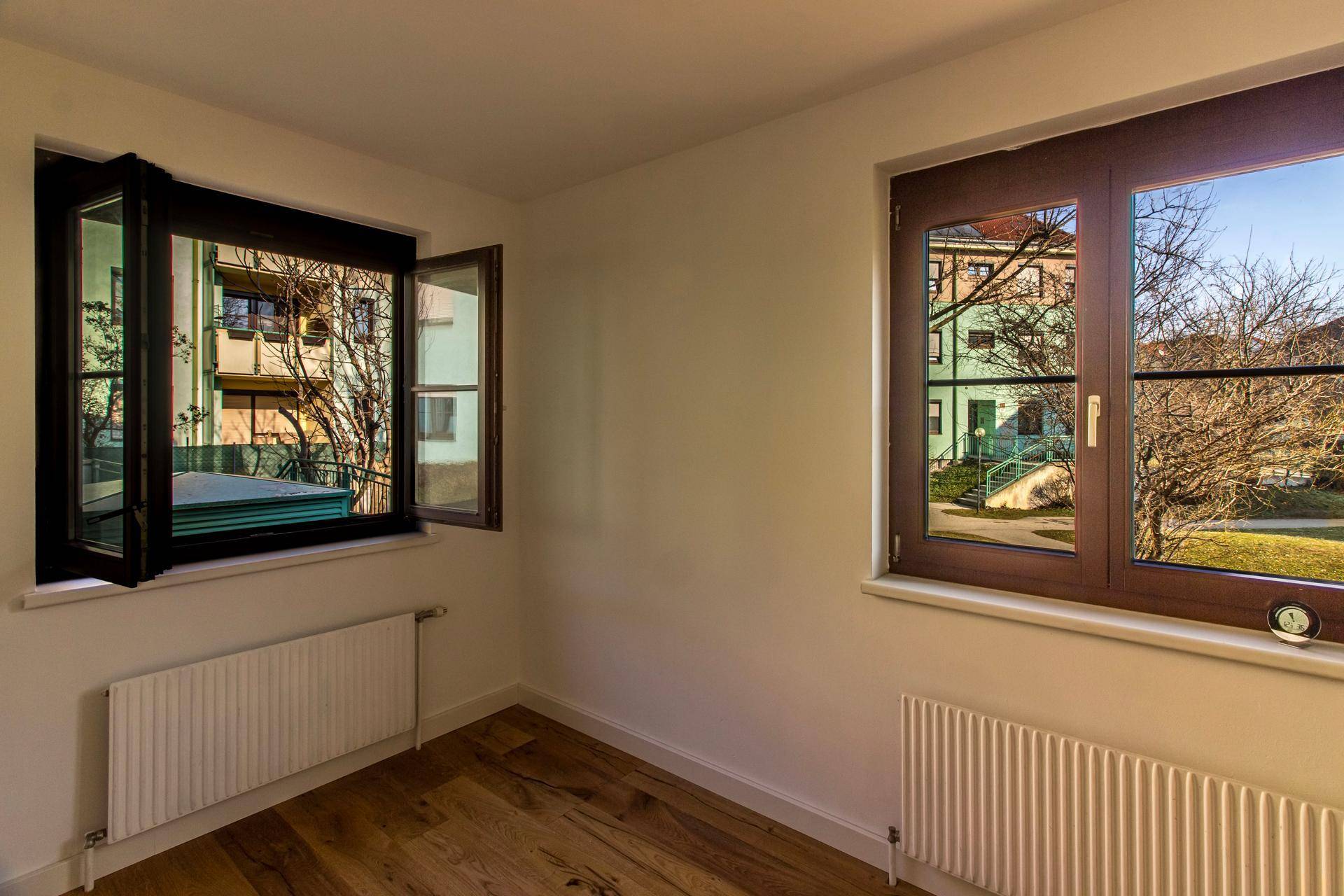 Helles Zimmer mit zwei Fenstern, Holzboden und Blick auf die grüne Umgebung.