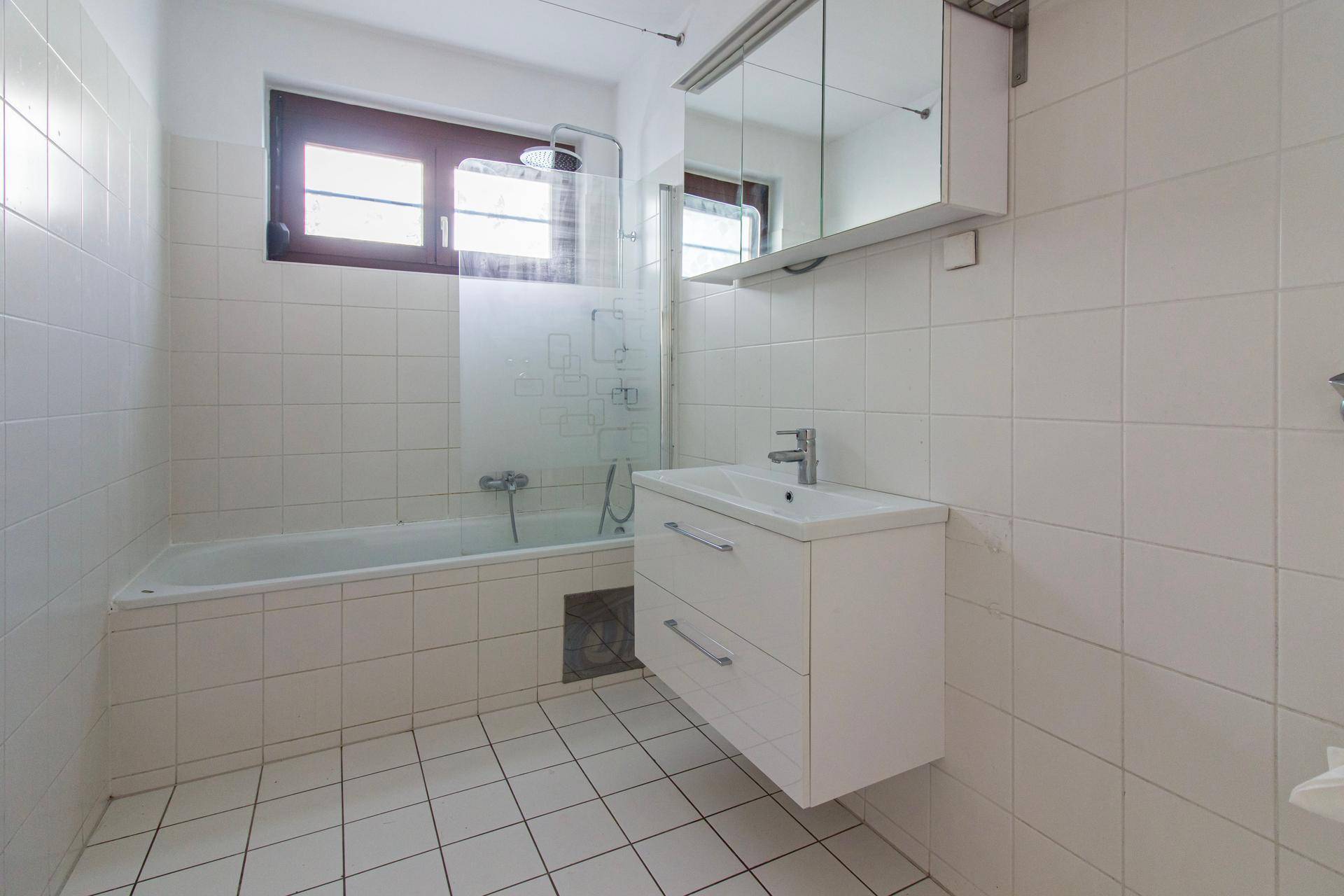 Badezimmer mit weißen Fliesen, Badewanne mit Duschabtrennung und Waschtisch mit Spiegelschrank.