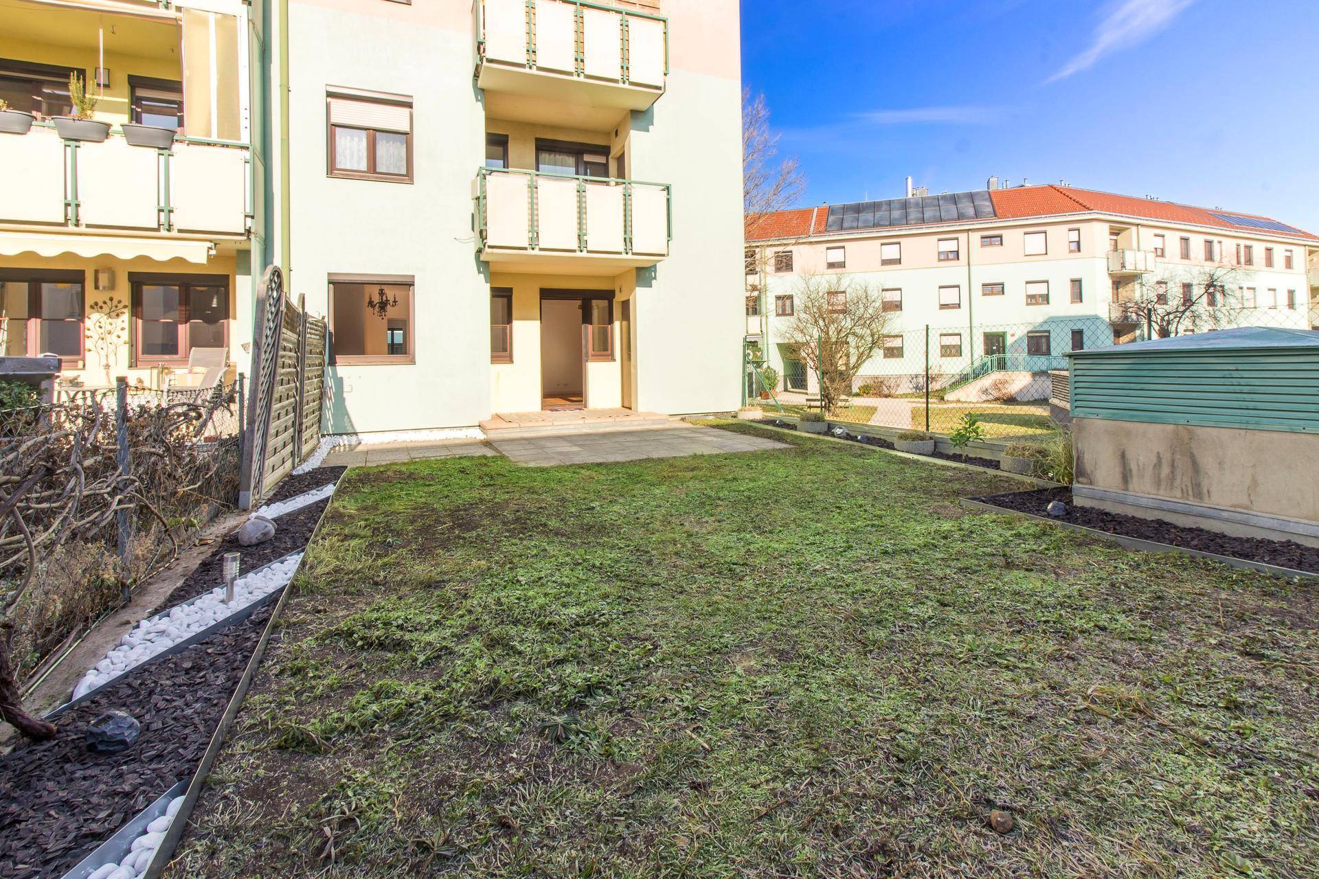 Garten mit Rasenfläche und gepflasterter Terrasse, umgeben von Wohngebäuden.