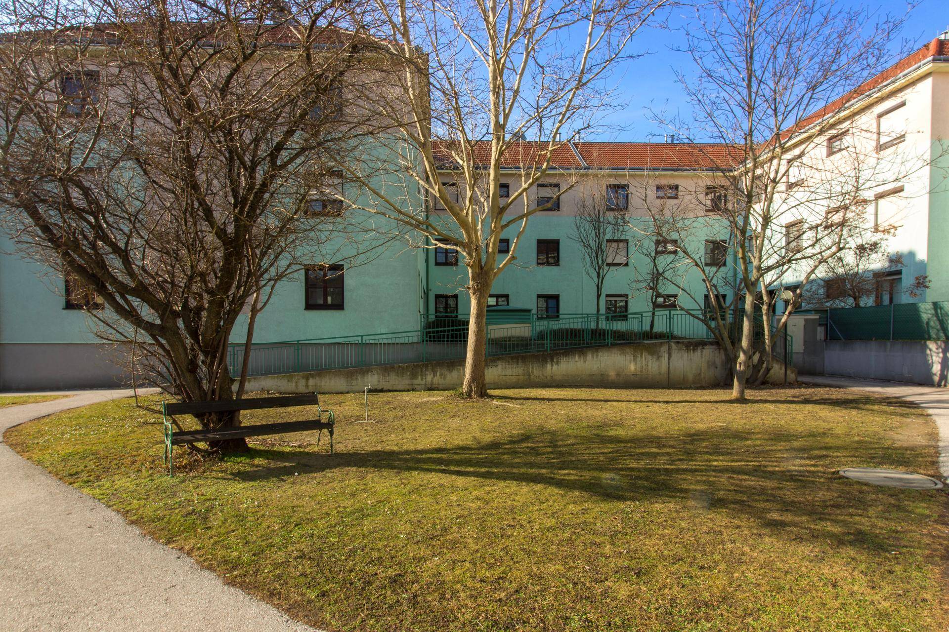 Außenbereich mit Grünfläche, Bäumen und einer Bank, umgeben von Wohngebäuden.