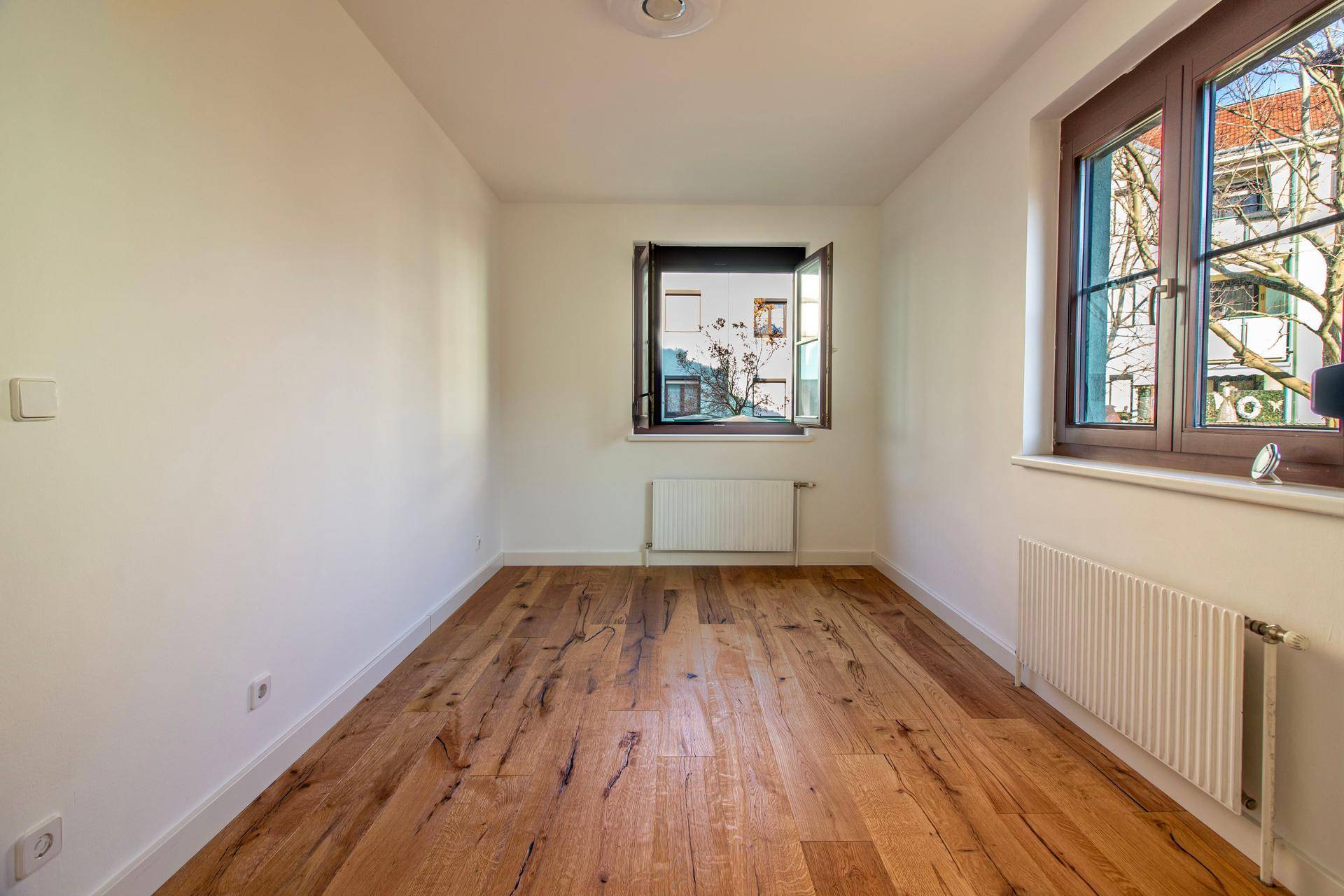 Langes, schmales Zimmer mit Holzboden und zwei Fenstern für viel Tageslicht.