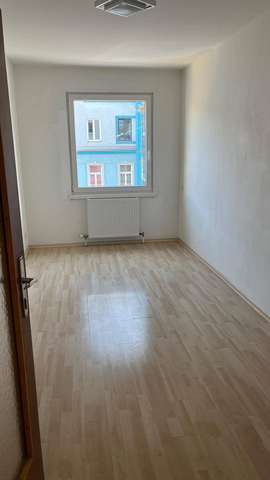 Leeres Schlafzimmer mit hellem Laminatboden, weißen Wänden und einem Fenster mit Stadtblick.
