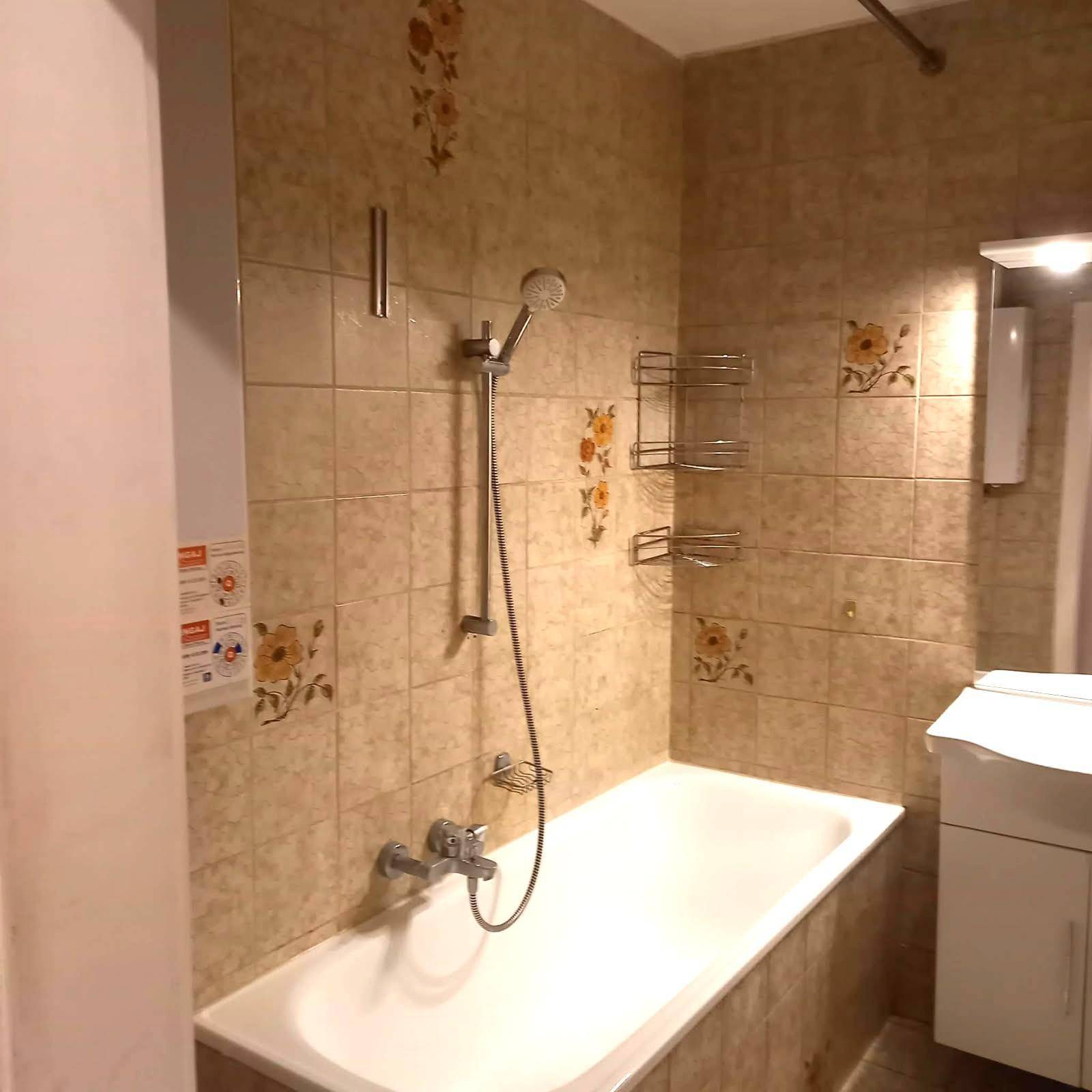 Badezimmer mit beigefarbenen Fliesen, Badewanne mit Duschfunktion und einem kleinen Waschbecken.