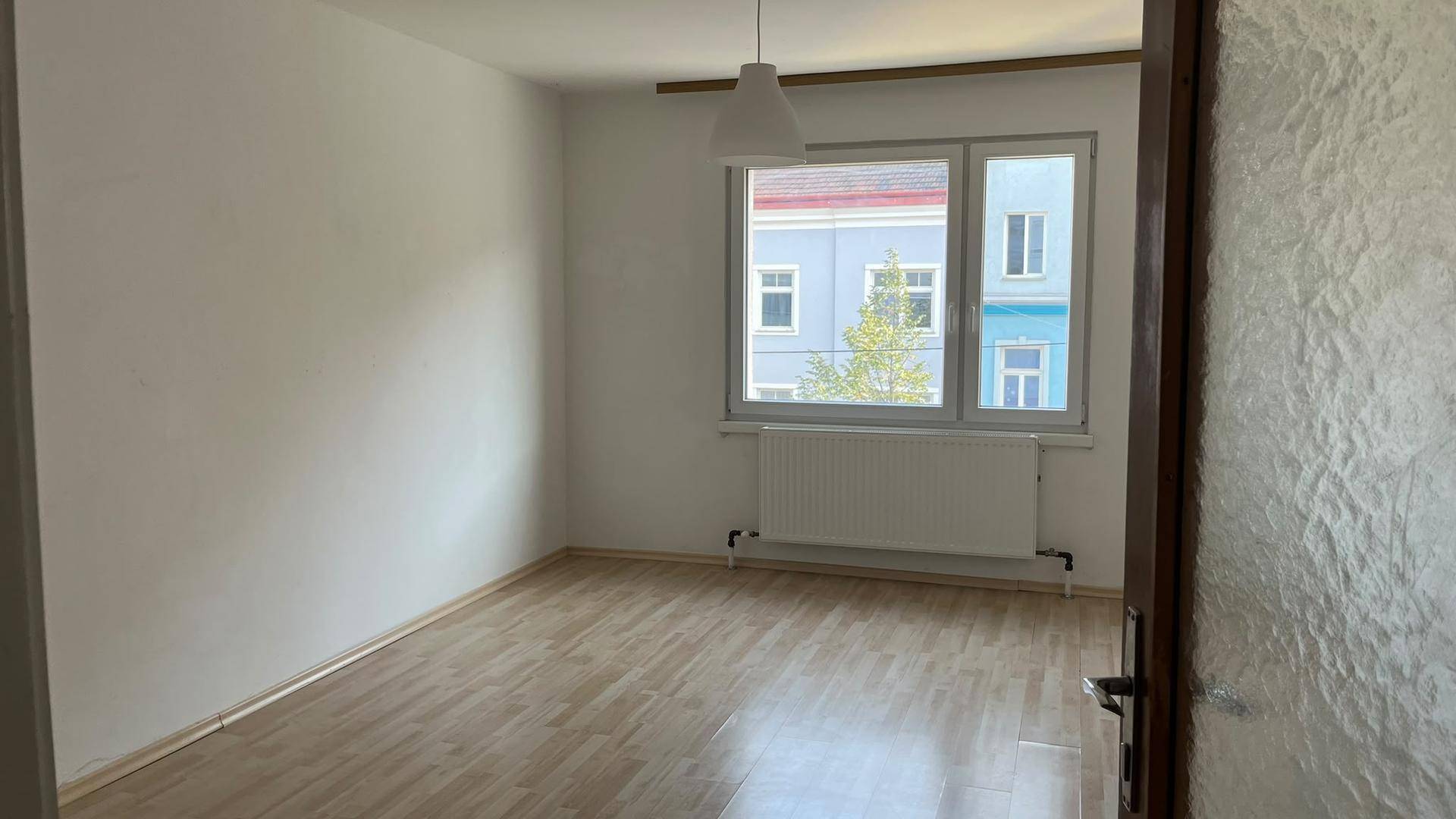 Geräumiges Zimmer mit hellem Laminatboden, weißen Wänden und einem großen Fenster.