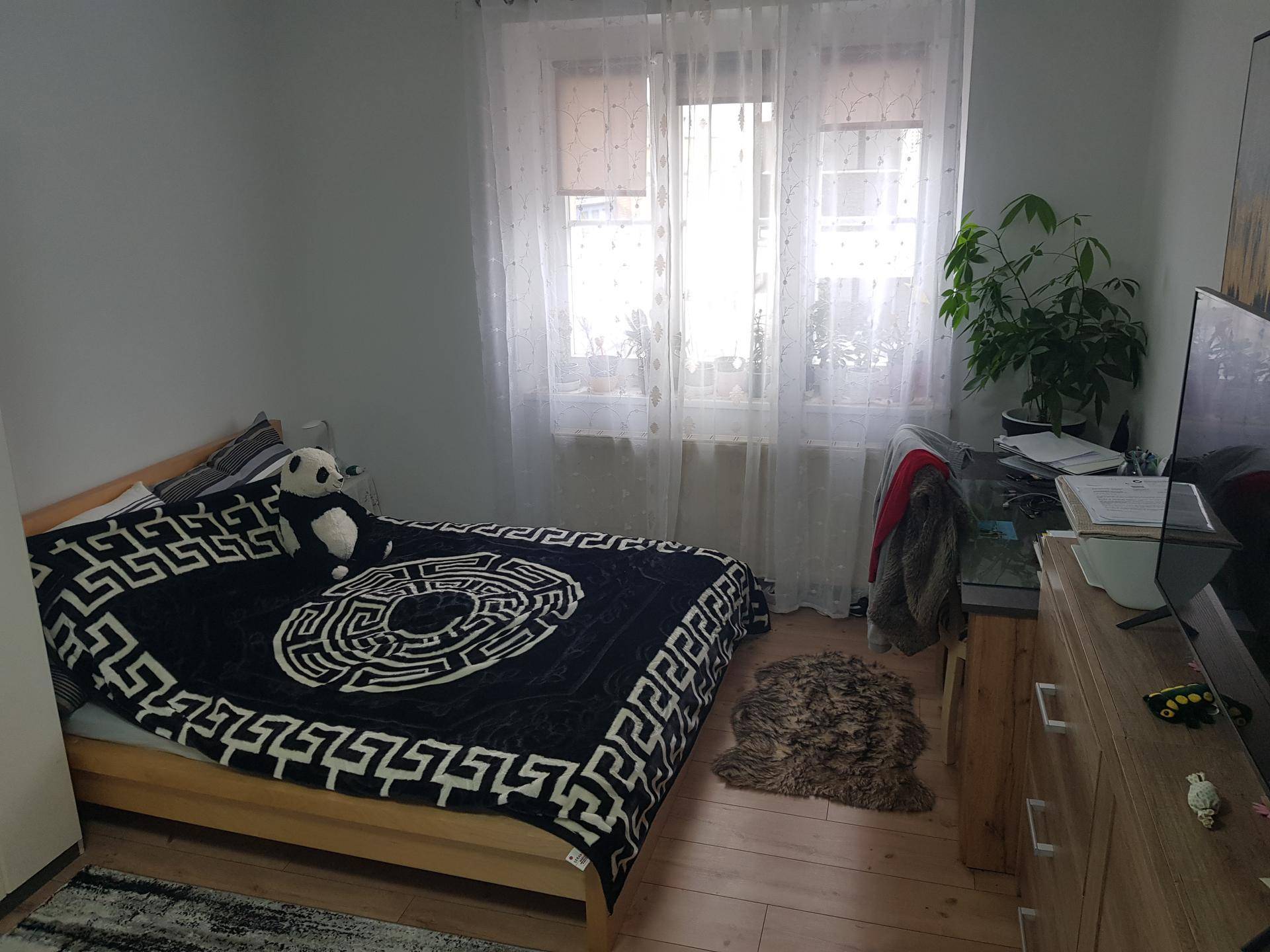 Helles Schlafzimmer mit Bett, Schreibtisch und Fenster mit transparenten Vorhängen.