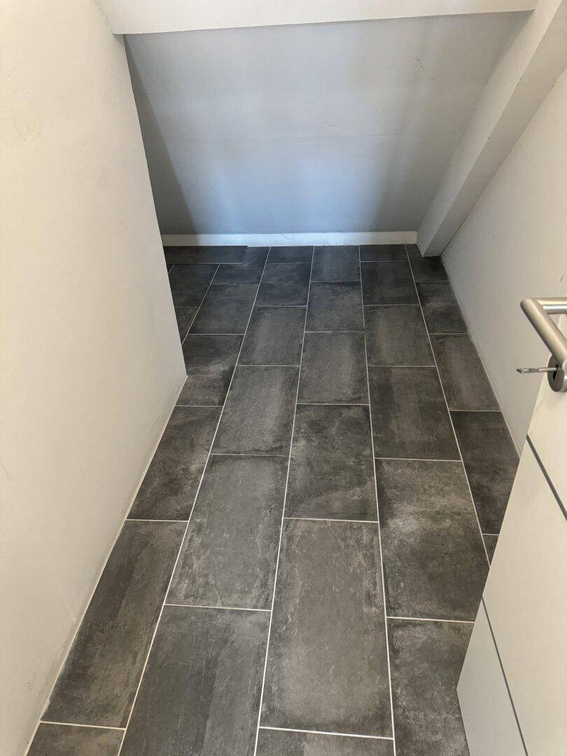 Schmaler Bereich unter einer Treppe mit dunklem Fliesenboden und hellen Wänden.