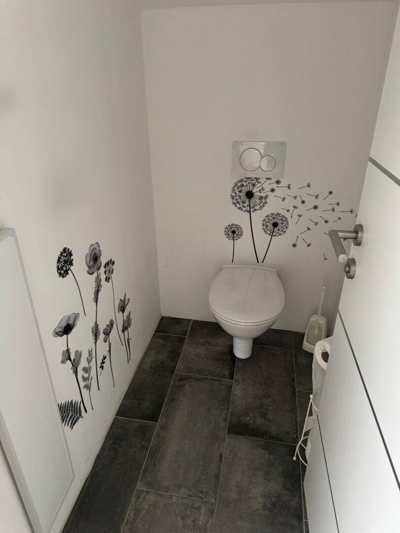 Gäste-WC mit wandhängender Toilette, dunklem Fliesenboden und dekorativen Wandstickern.