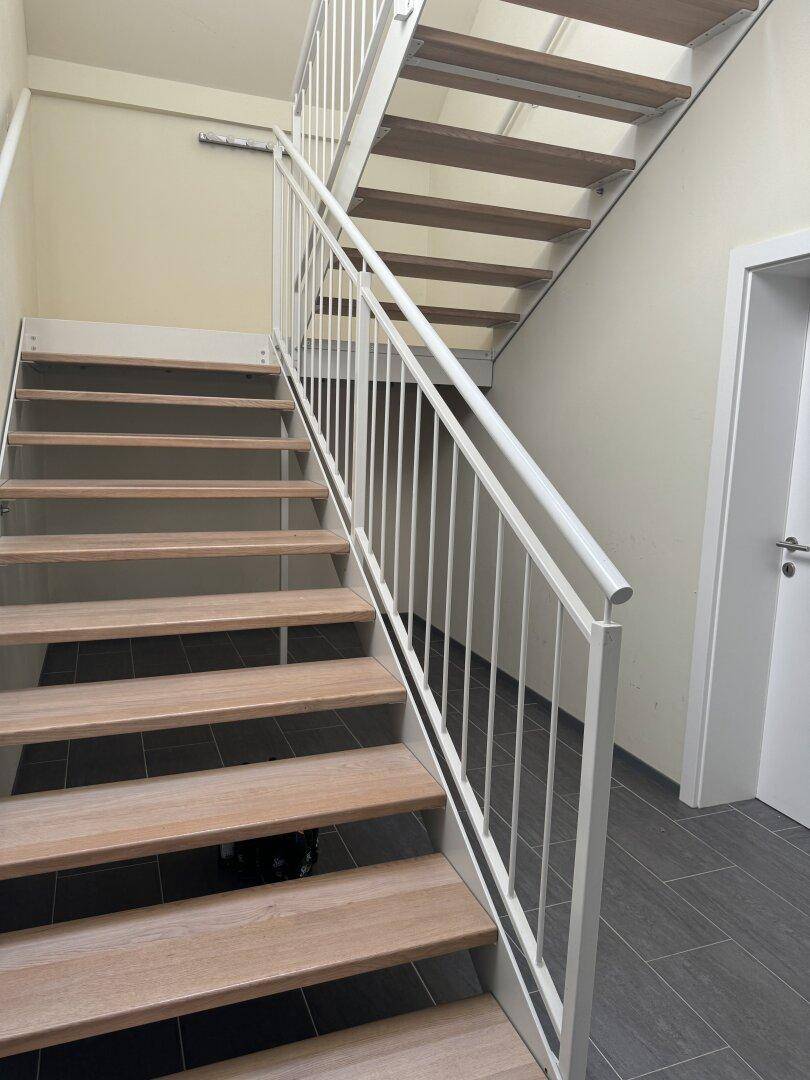 Moderne Holztreppe mit weißem Metallgeländer und Fliesenboden im Eingangsbereich.