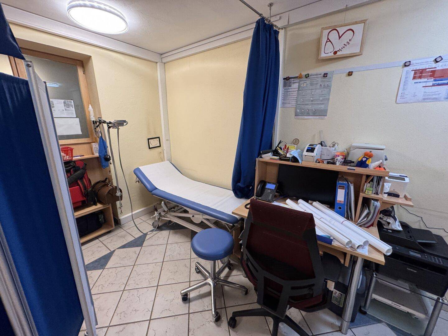 Untersuchungszimmer mit Behandlungsliege, Schreibtisch und hellen Fliesen.