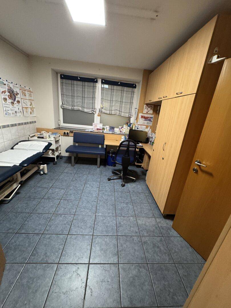 Untersuchungszimmer mit Behandlungsliege, Schreibtisch und dunklen Fliesen.