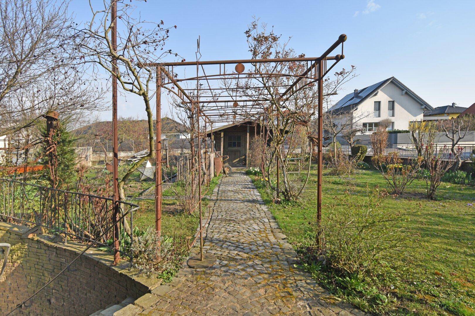 Gartenweg aus Kopfsteinpflaster führt zu einem Gartenhaus unter einer Pergola.