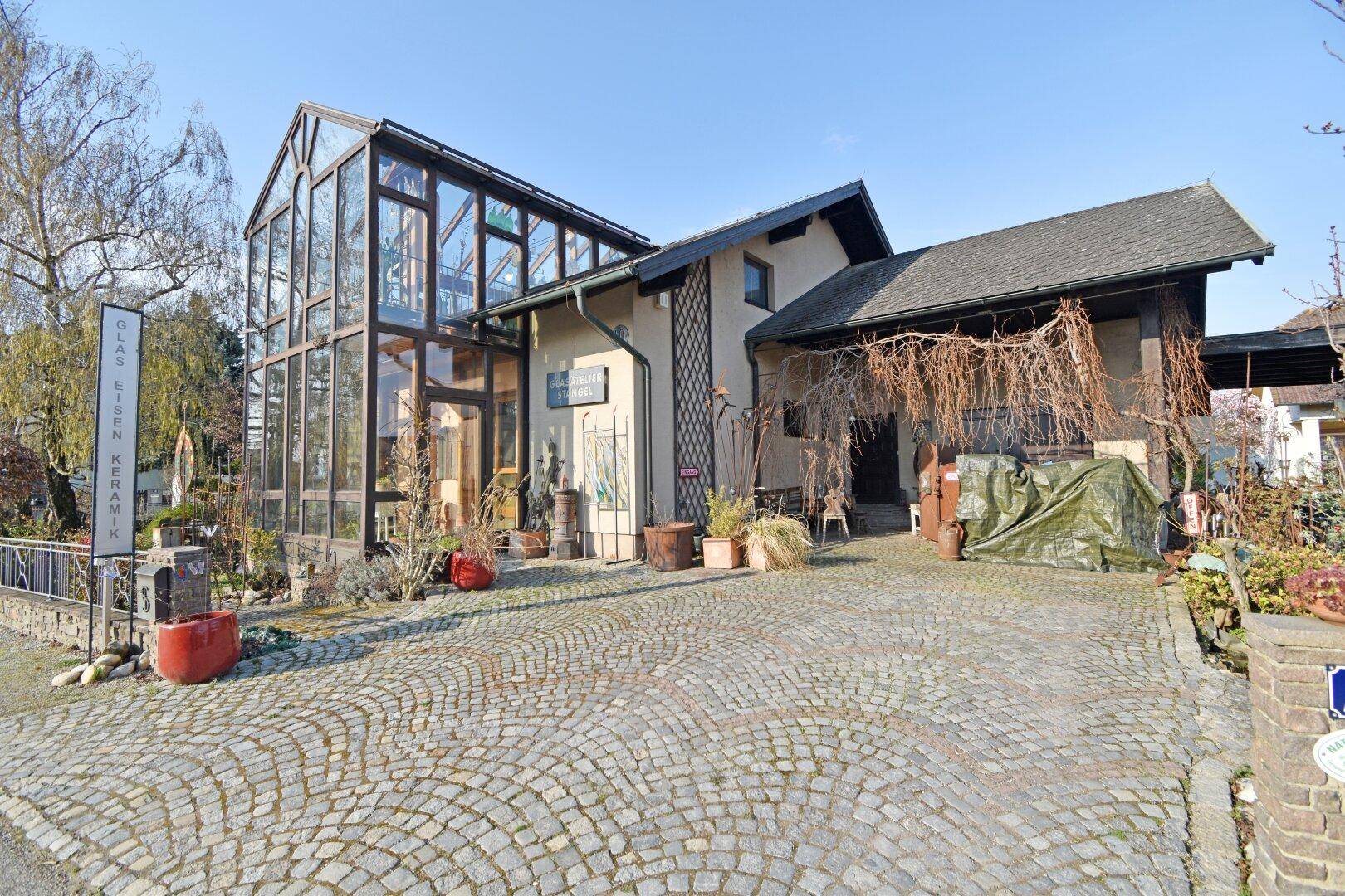 Frontansicht des Hauses mit großem Wintergarten, gepflasterter Einfahrt und Schild.