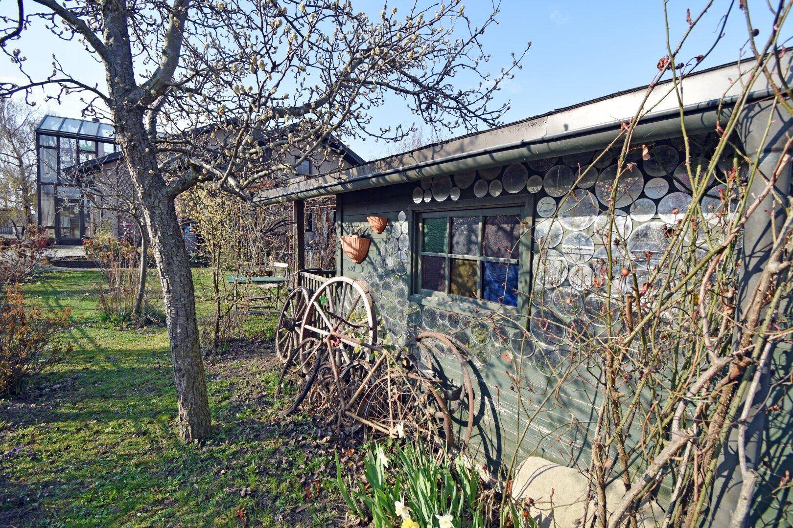 Rustikales Gartenhaus mit dekorativen Fenstern und alten Wagenrädern im Garten.