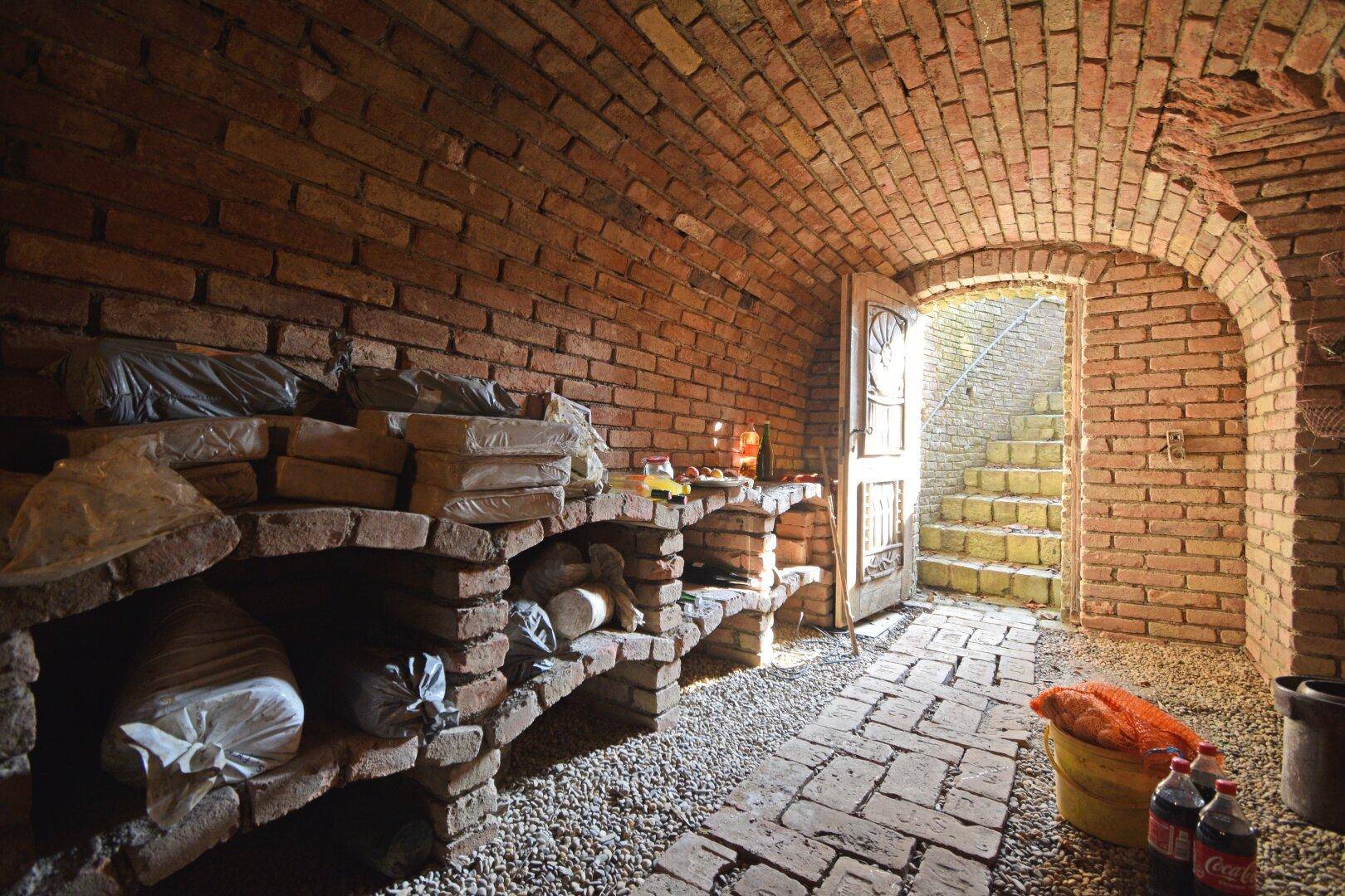 Historischer Gewölbekeller aus Ziegelstein mit Lagerregalen und Kiesboden.