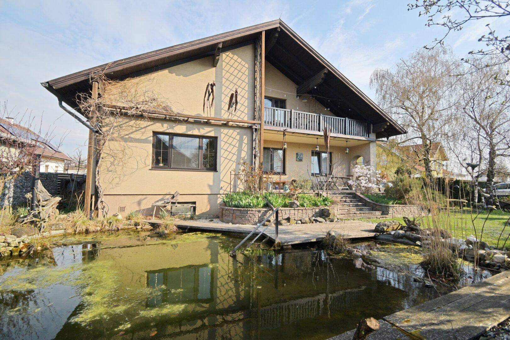 Hausfassade mit Balkon und Teich im Garten, umgeben von Grünflächen und Bäumen.