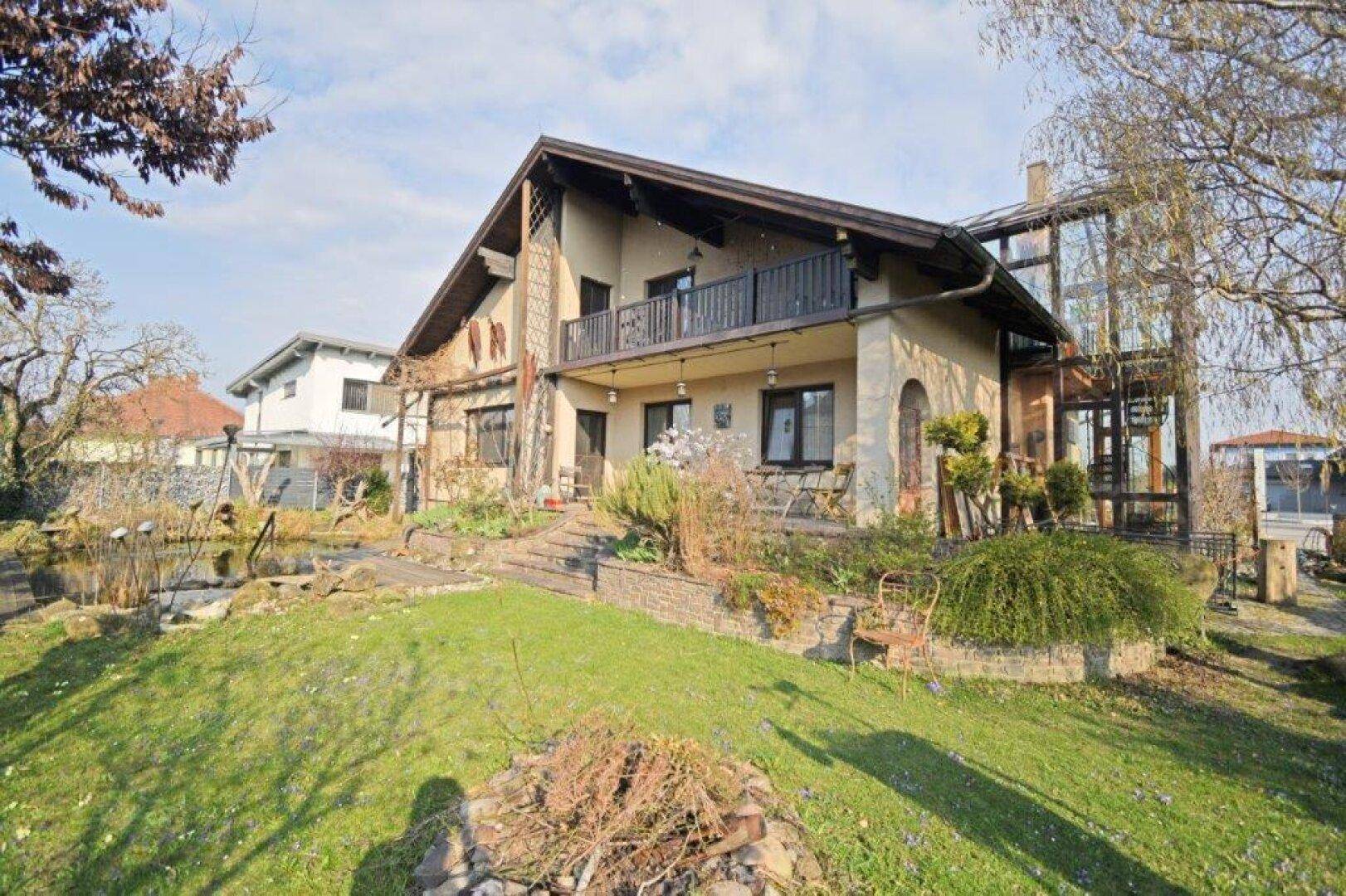 Hausfassade mit Balkon und Teich im Garten, umgeben von Grünflächen und Bäumen.