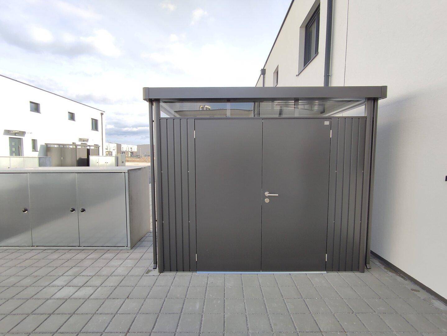 Moderner Abstellraum oder Carport mit dunklen Türen und heller Fassade des angrenzenden Gebäudes.