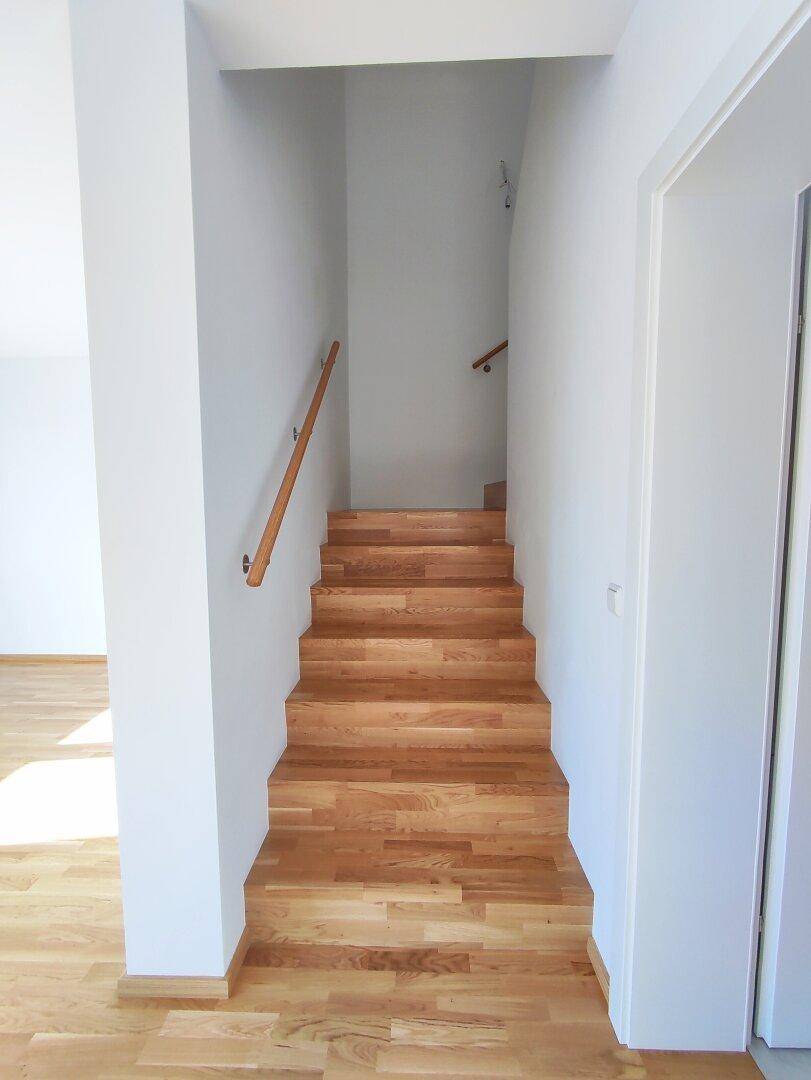 Holztreppe mit hellem Geländer führt in die oberen Stockwerke, umgeben von weißen Wänden.