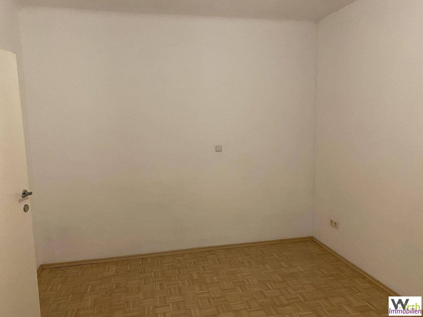 Schlichter, leerer Raum mit Parkettboden und weißen Wänden.