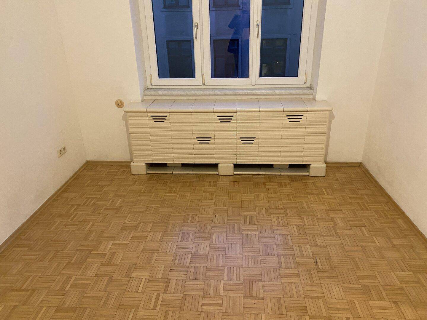 Heller Raum mit Parkettboden und Fenster, darunter ein verkleideter Heizkörper.