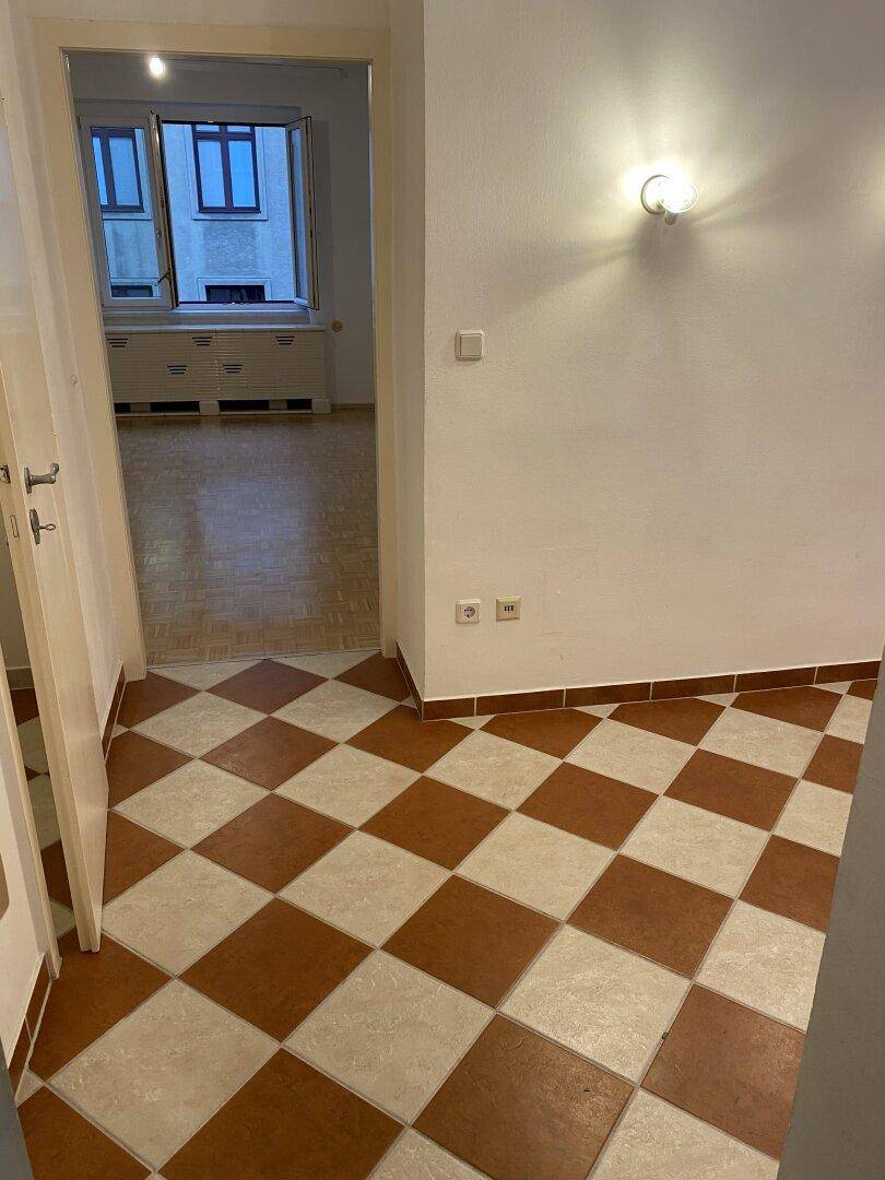 Flur mit Schachbrettmuster-Fliesen, der zu einem angrenzenden Raum mit Parkettboden führt.