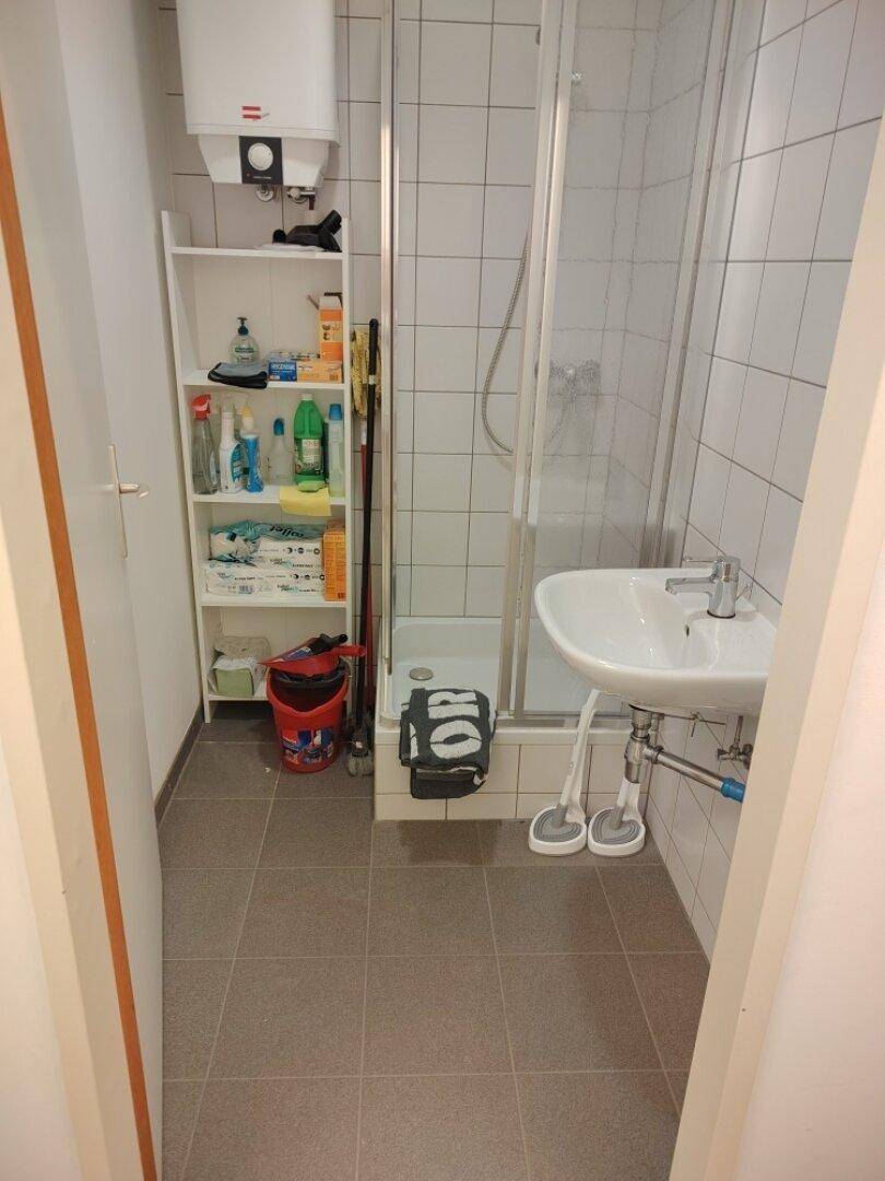 Kleines Badezimmer mit Duschkabine, Waschbecken und Warmwasserboiler, funktional eingerichtet.