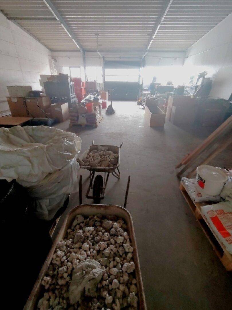 Blick durch die Lagerhalle mit Material auf Paletten und Schubkarren, hell erleuchtet am Ende.