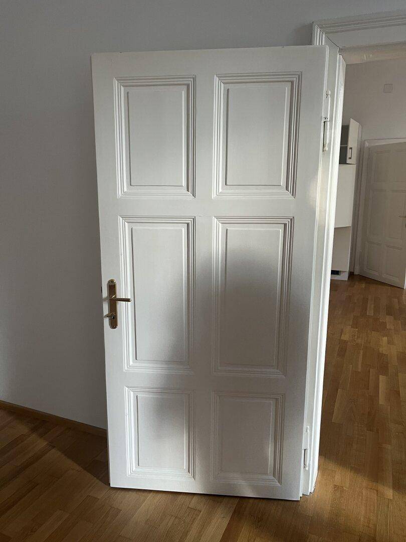 Weiße Holztür mit traditionellem Design und Messinggriff, in einem hellen Innenraum.