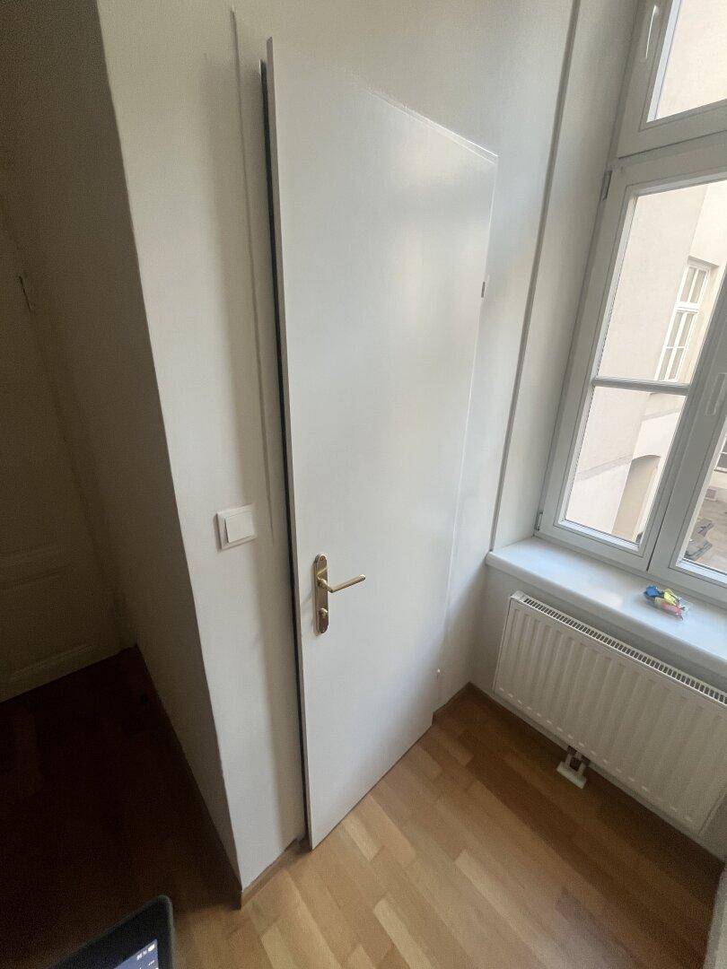 Schlichte weiße Tür neben einem Fenster mit Heizkörper, in einem hellen Raum.