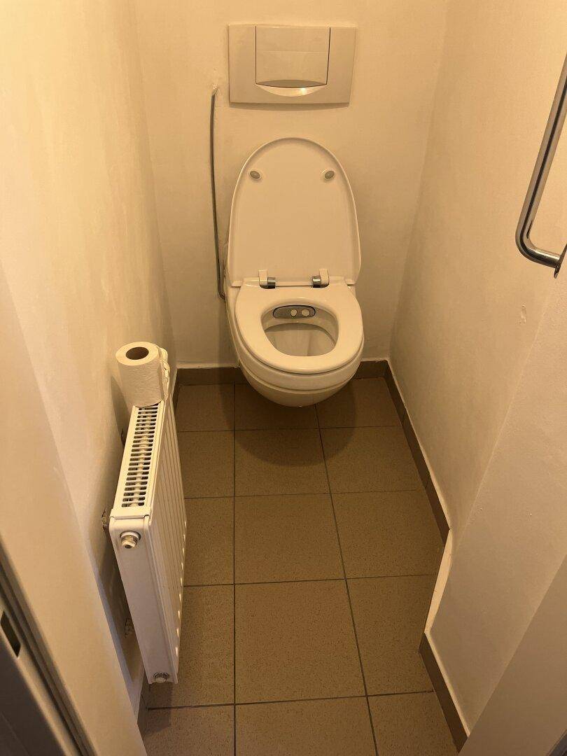 Kleines Gäste-WC mit Wand-WC, Heizkörper und braunen Bodenfliesen, funktional eingerichtet.