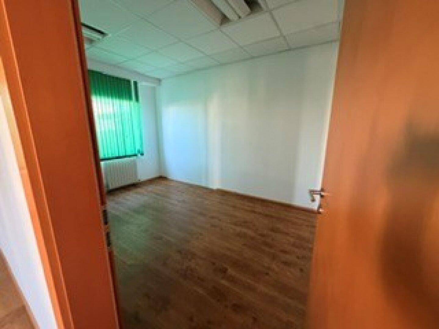Leeres Büro mit Laminatboden, weißen Wänden und grünen Jalousien vor dem Fenster.