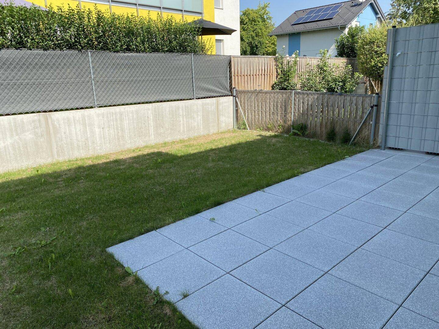Gepflegte Rasenfläche neben einer gepflasterten Terrasse, umgeben von einem Zaun und Hecken.